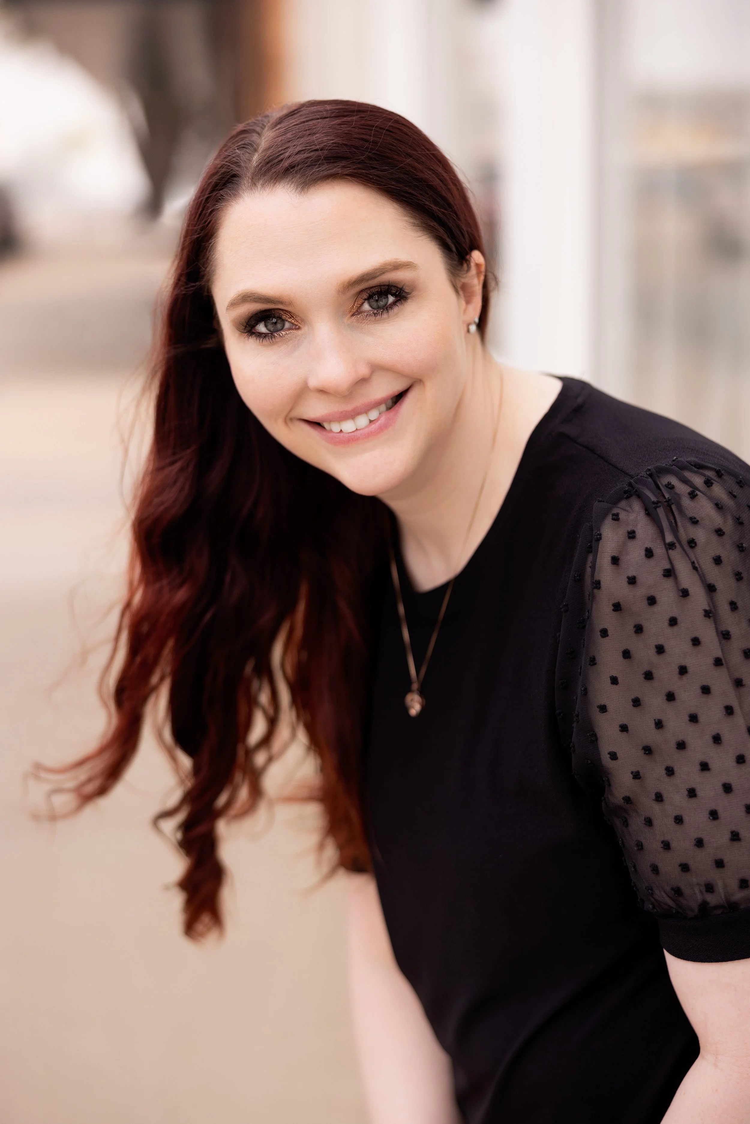 A woman with long, wavy red hair is smiling at the camera. She is wearing a black top with sheer, polka-dotted sleeves and a necklace. The background is blurred, suggesting an indoor setting with natural light.