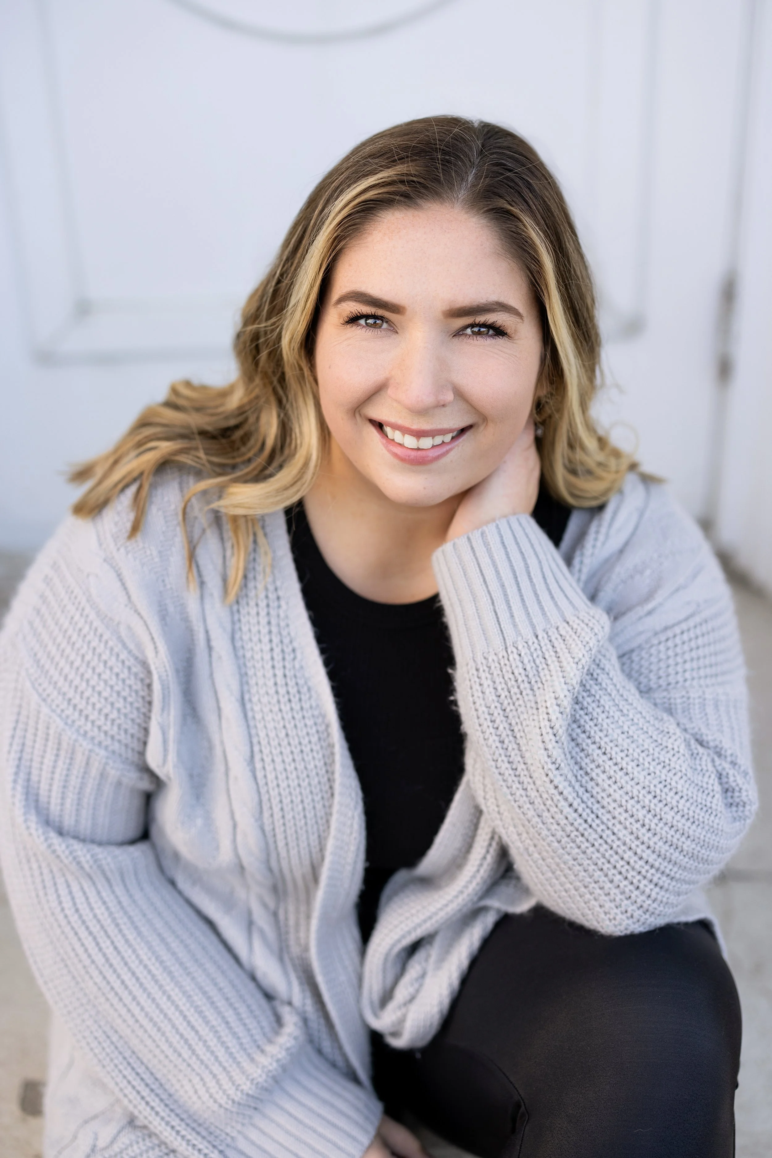 A woman with long blonde hair and fair skin, smiling and looking at the camera. She is wearing a gray knit sweater and a black top, sitting against a light-colored wall.
