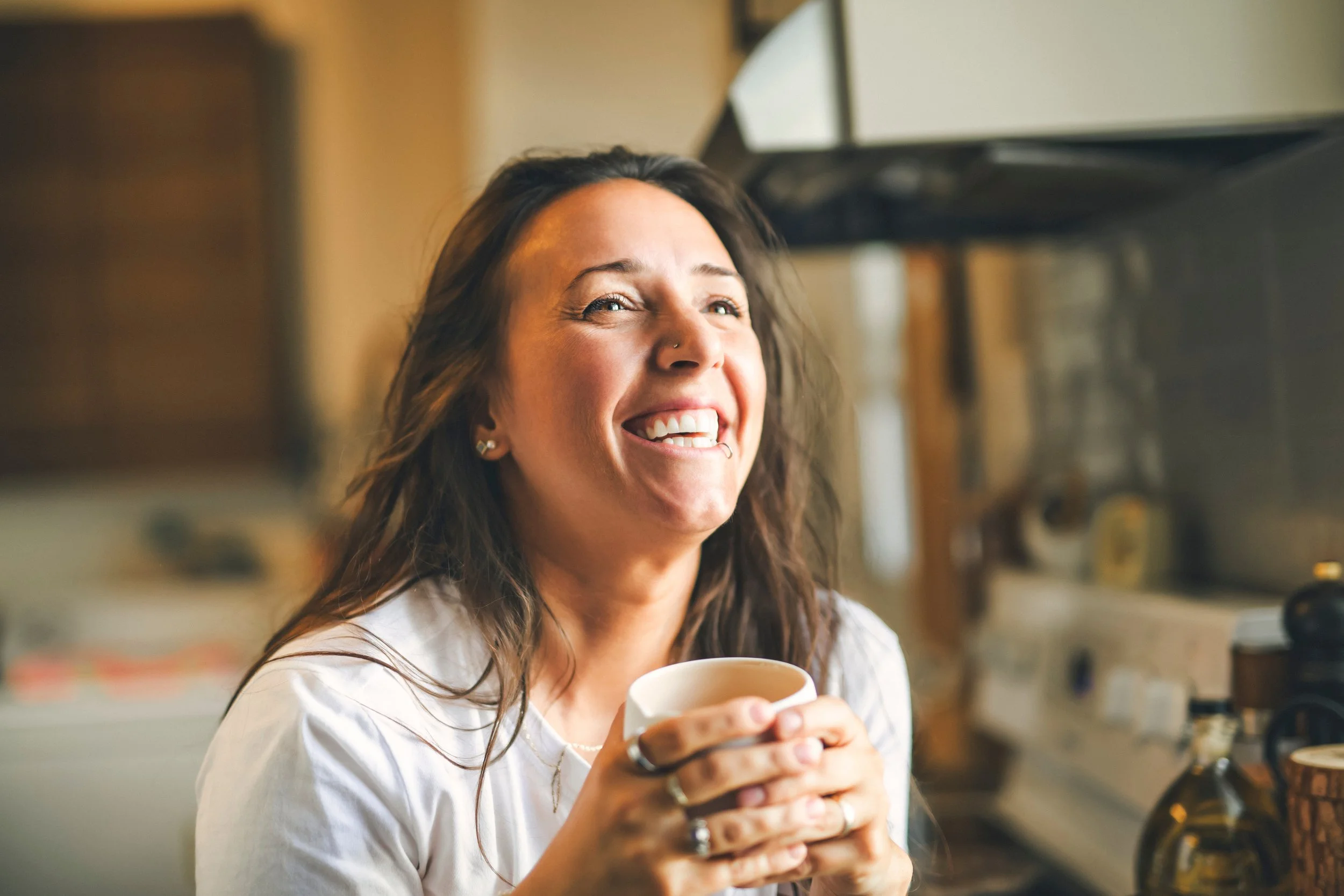 A woman with long brown hair smiling and holding a white mug in a cozy kitchen.
