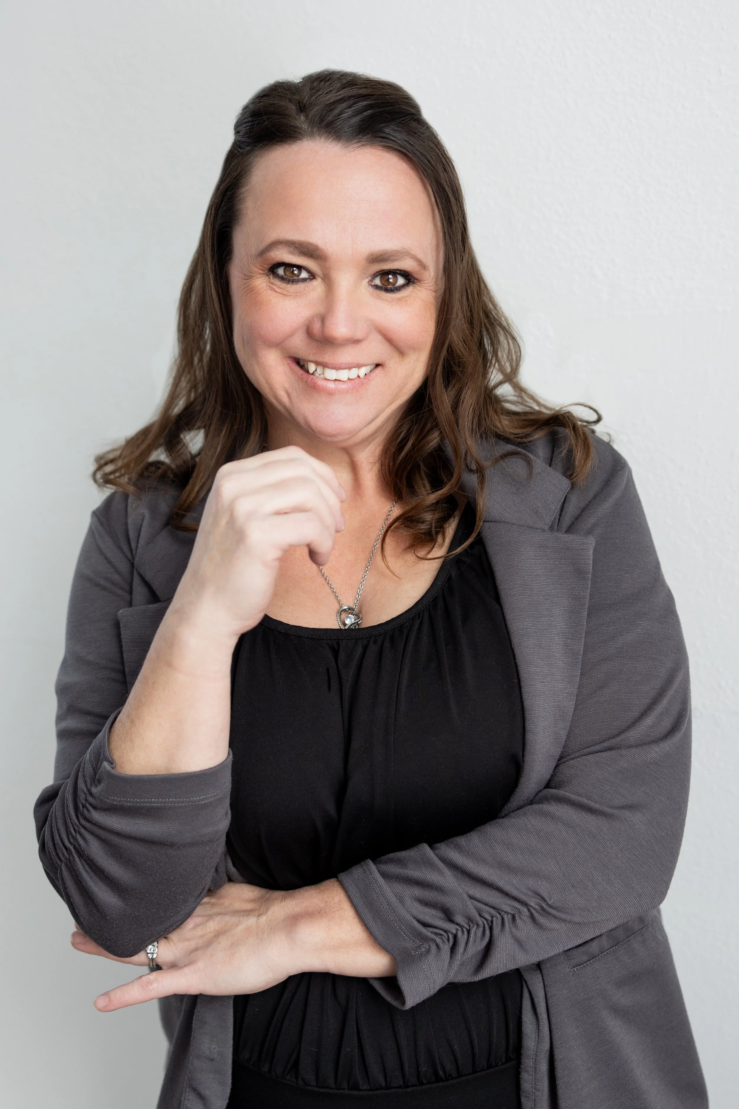 A woman with brown hair and brown eyes smiling, wearing a gray blazer over a black top, with her left arm crossed and right hand touching her chin, standing against a plain white background.