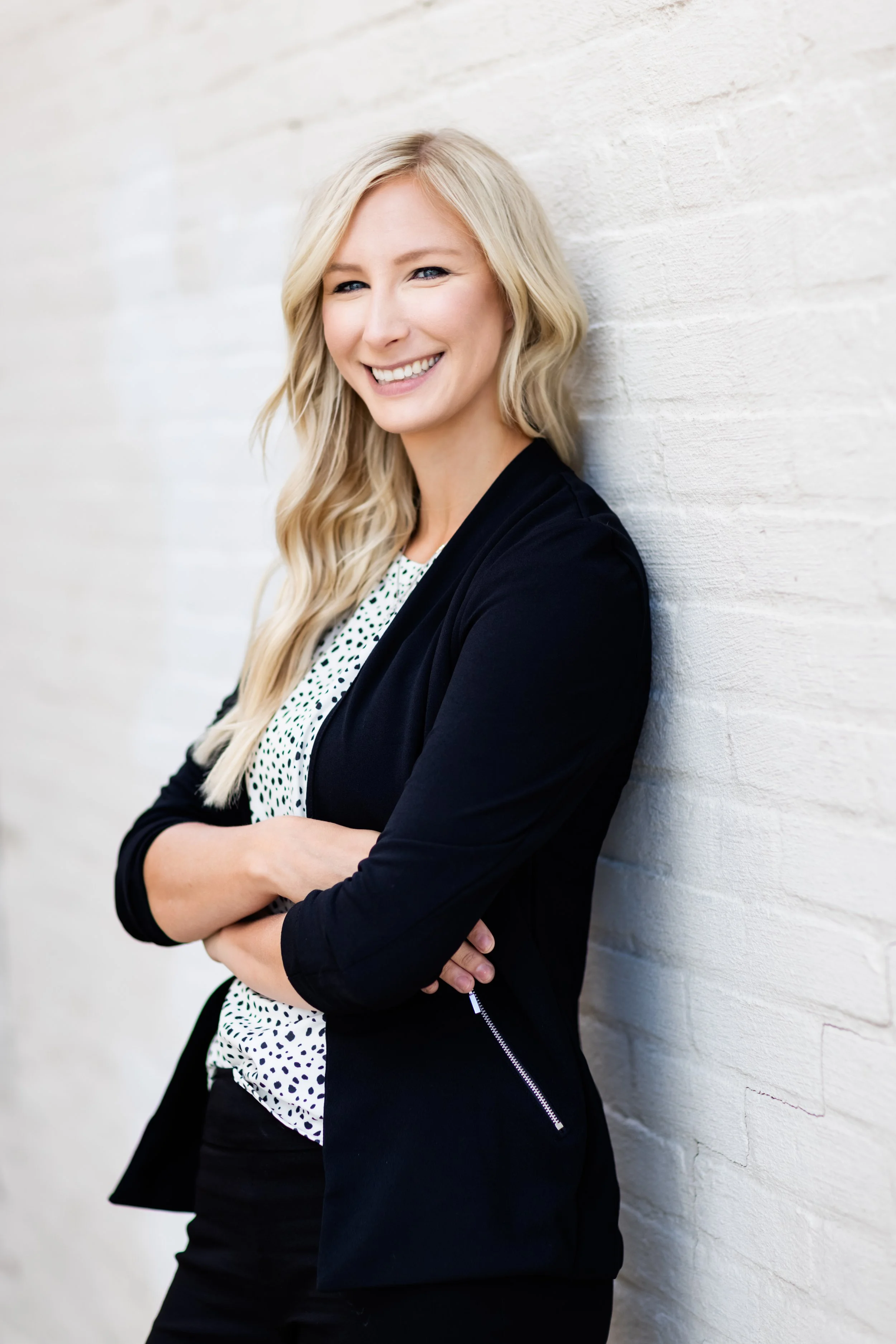 A smiling blonde woman in a polka dot blouse and black blazer is standing with her back against a white brick wall, arms crossed.