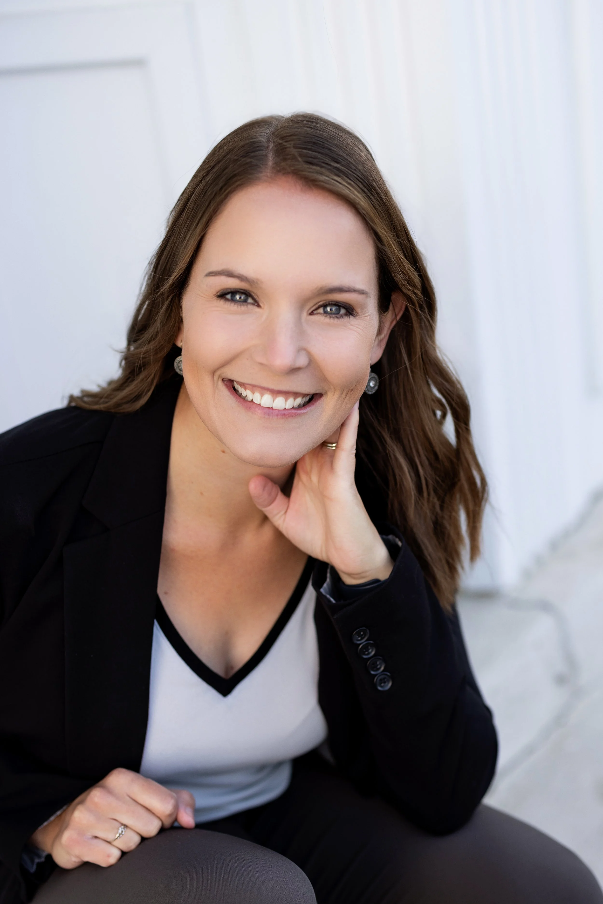 A woman with long brown hair smiling, sitting in front of a white wooden wall, wearing a black blazer and a white shirt with black trim.