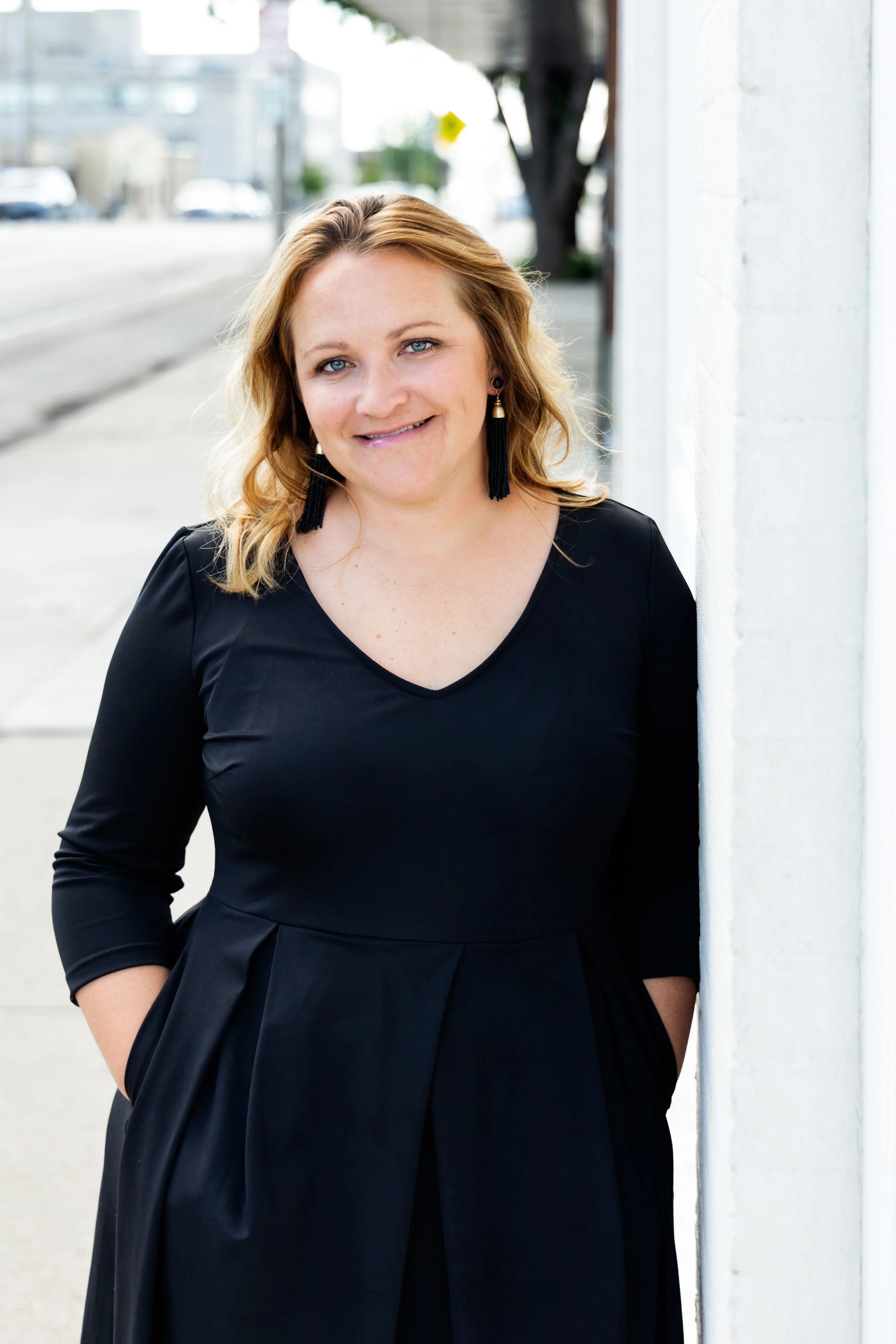 A woman with blonde hair and blue eyes is smiling, wearing a black dress with tassel earrings, and standing outdoors on a sidewalk next to a white wall, with blurred buildings and trees in the background.
