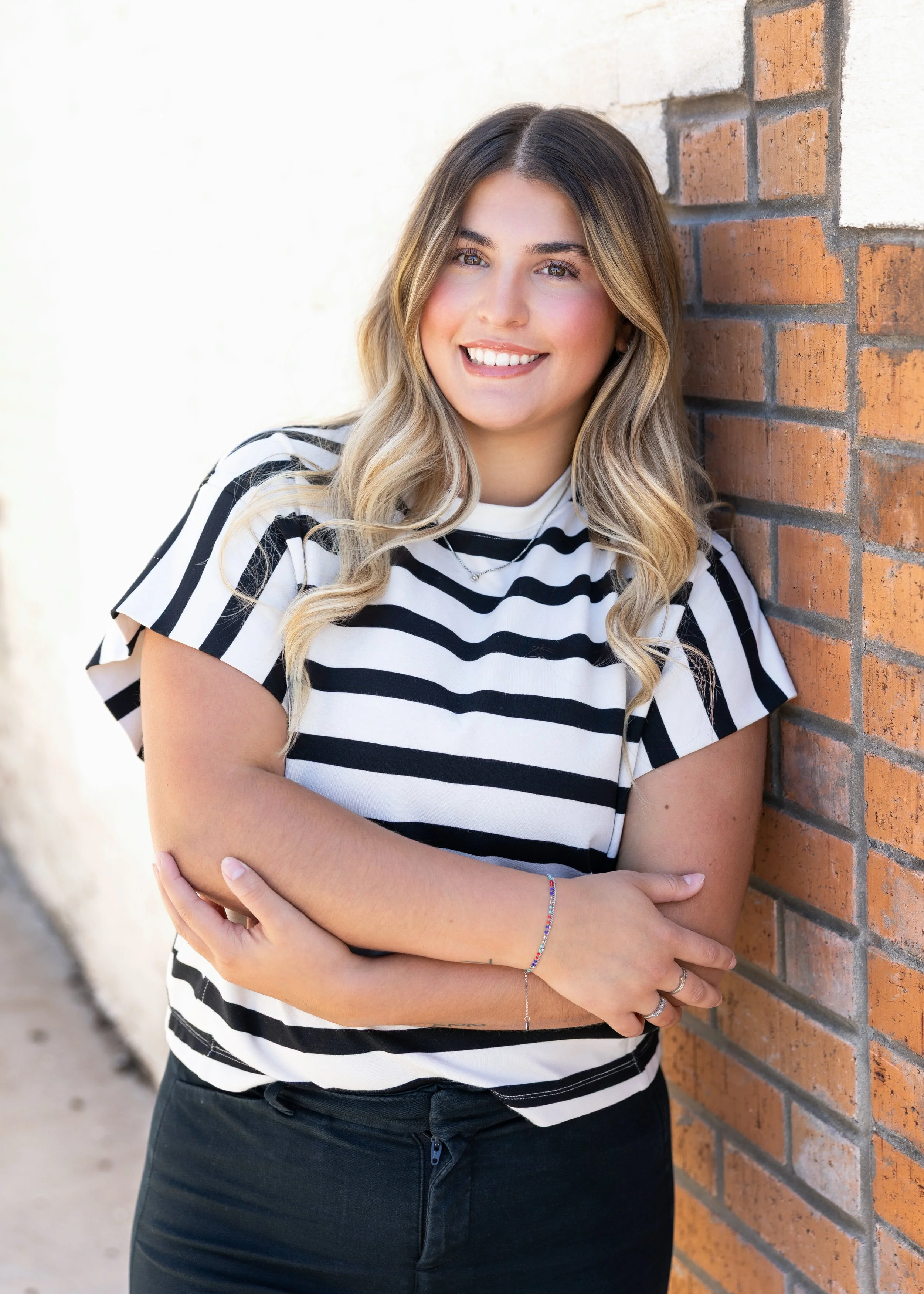 A young woman with long, wavy blonde hair, smiling, leaning against a brick wall with her arms crossed in front of her. She is wearing a black and white striped t-shirt and black pants.