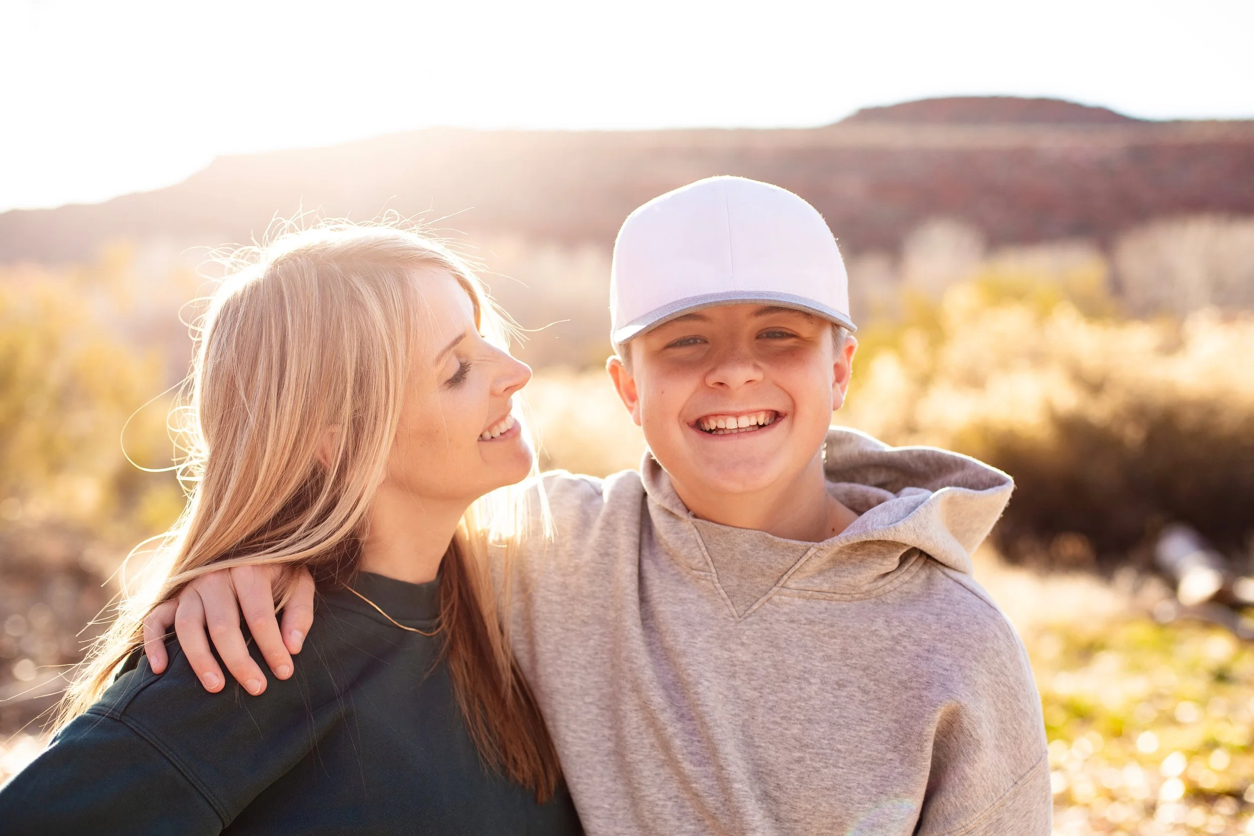 A woman with long blonde hair and a man with a white cap are smiling and embracing outdoors in a sunny, autumn landscape.