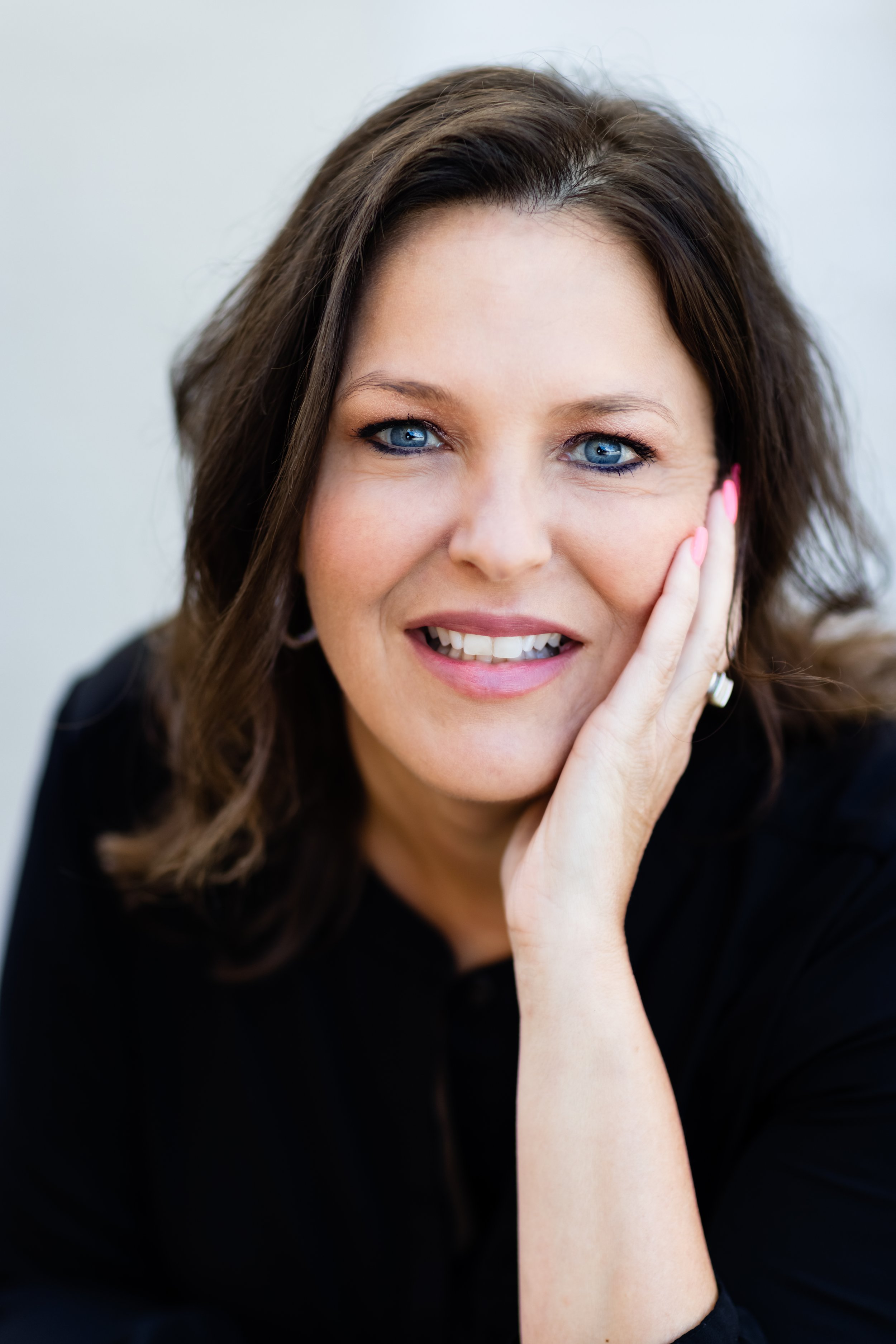 Close-up of a woman with wavy brown hair and bright blue eyes, smiling and touching her face with her right hand, wearing a black top and pink nail polish.
