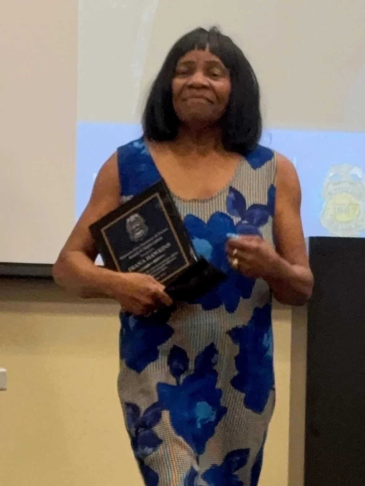 Woman in a blue and beige floral dress holding an award, standing in front of a white screen with a logo.