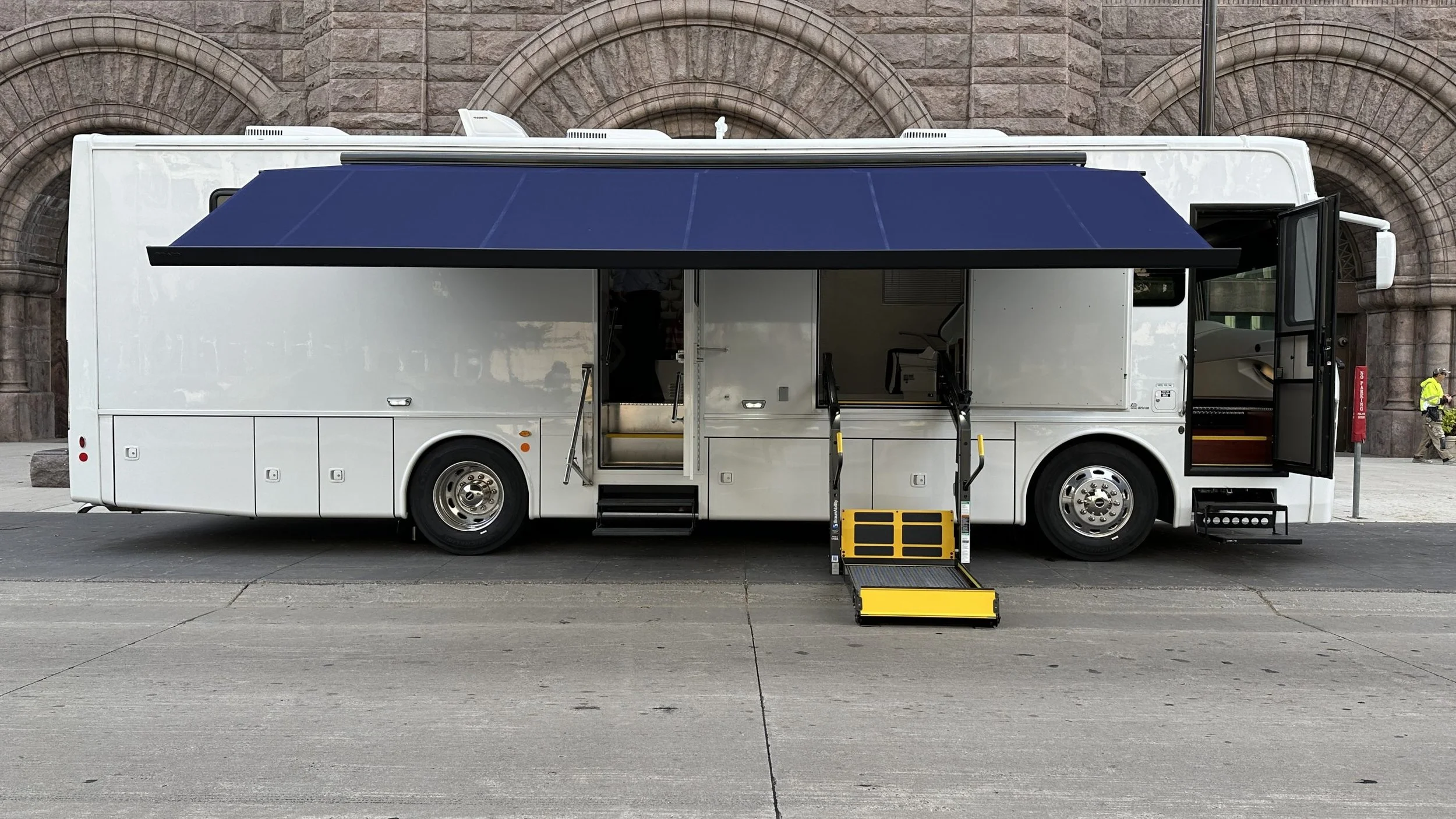 A white mobile tour bus with a blue retractable awning extends over the sidewalk, with foldable yellow and black steps at the entrance, parked in front of a stone building with arched windows.