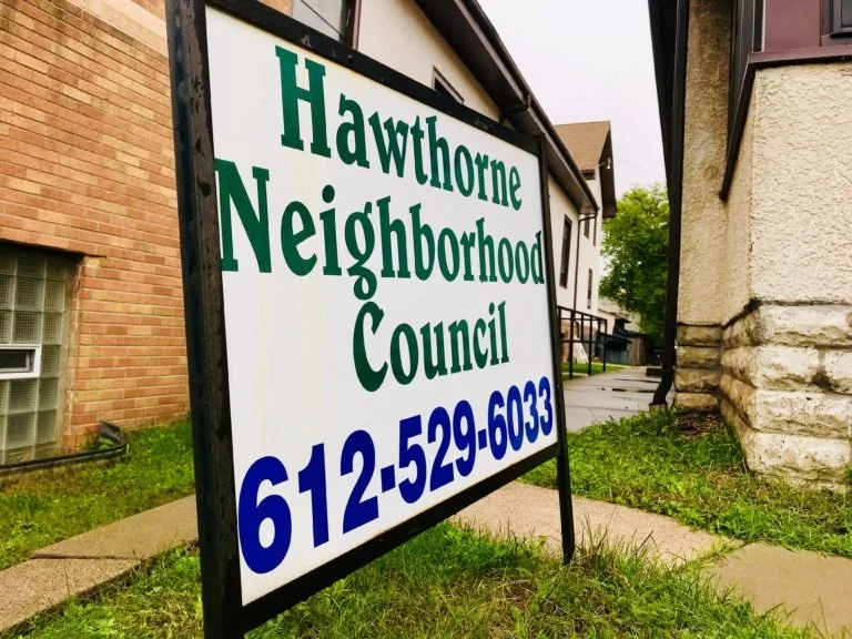 Signboard for Hawthorne Neighborhood Council with a phone number 612-529-6033 outside on a grassy area near buildings.