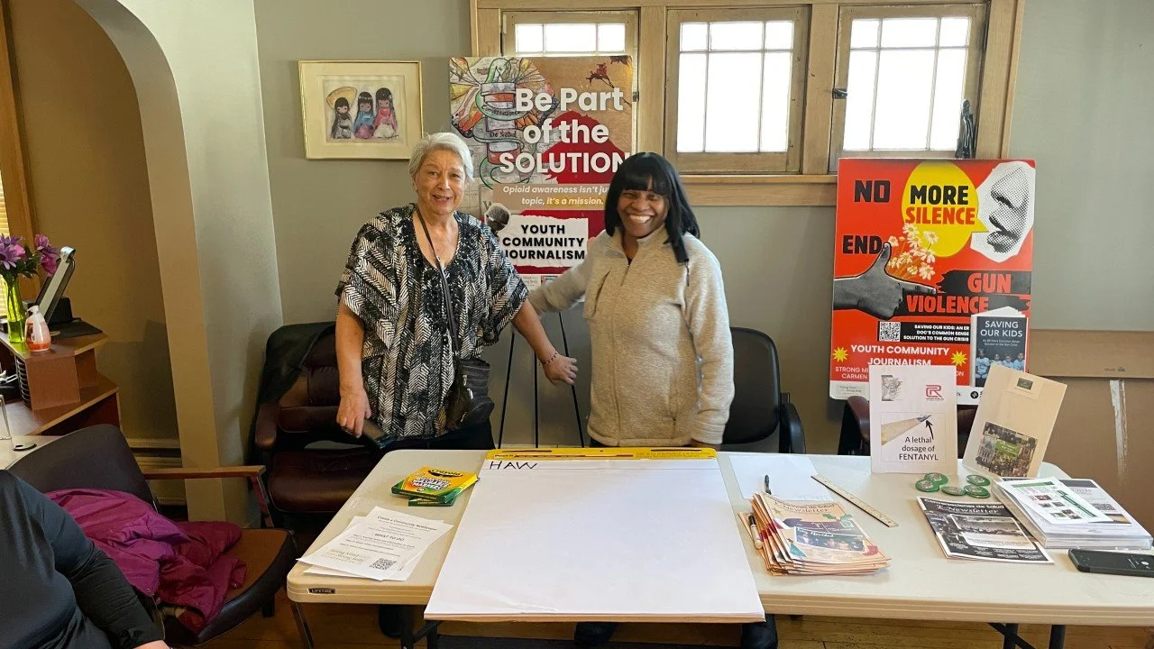 Two women smiling and standing at a table during a community event about youth and gun violence prevention, with informational posters and brochures on the table.