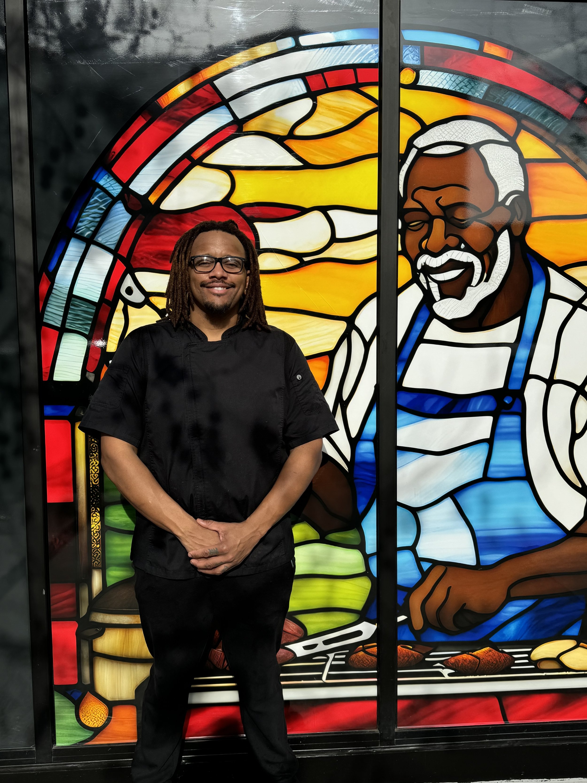 A man in a black chef's coat standing in front of a colorful stained glass window depicting a smiling man, vegetables, and a sunset. The window has bold outlines and bright colors.