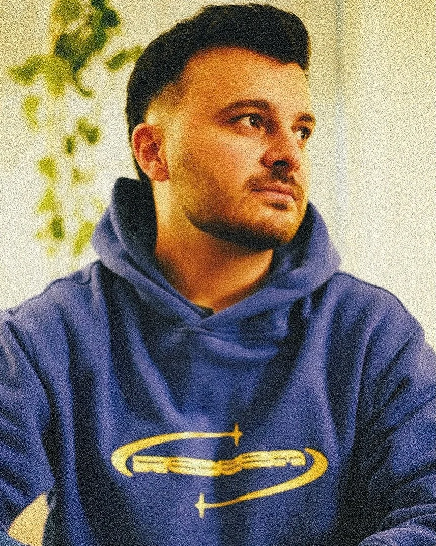 A young man with dark hair and a beard wearing a blue hoodie with yellow graphics, sitting indoors with a plant in the background.