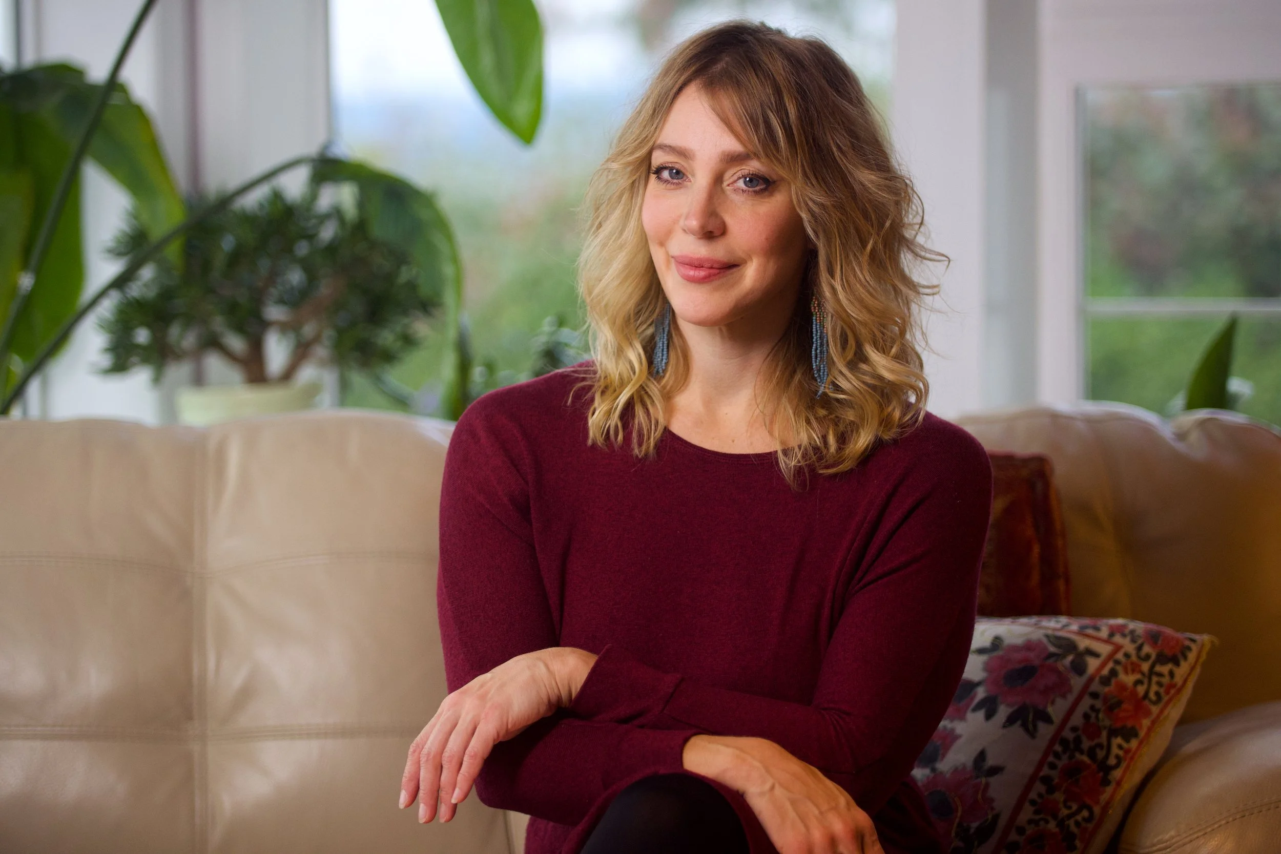 A woman with blonde, curly hair wearing a burgundy long-sleeve top, sitting on a beige sofa with a floral pillow in a well-lit room with large windows and green plants in the background.