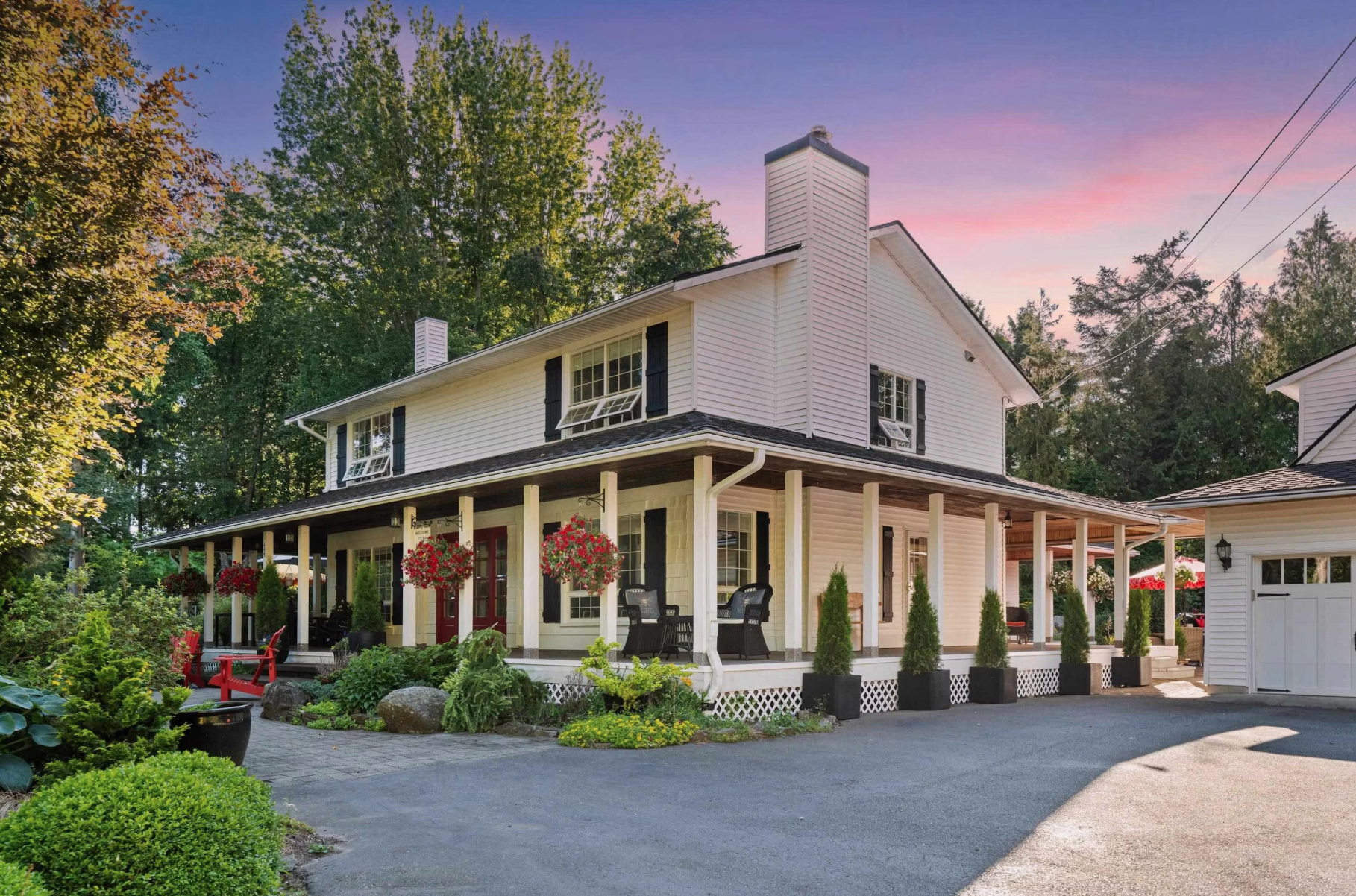 White two-story house with black shutters, porch with hanging flower baskets, potted plants, and outdoor furniture, surrounded by trees and greenery at sunset.