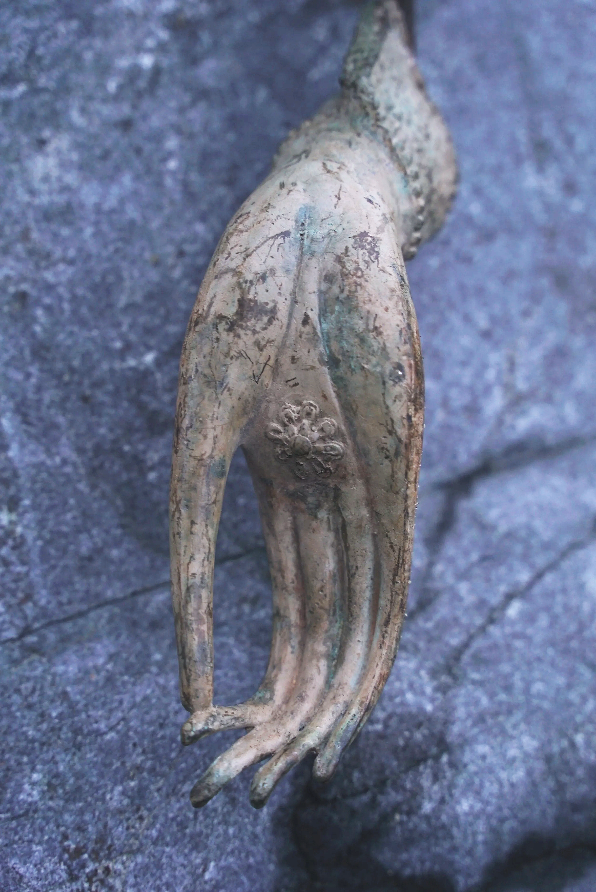 Close-up of an ornate, antique metal hand sculpture with detailed fingers, a floral decoration on the wrist, and a textured weathered surface, resting on a stone surface.