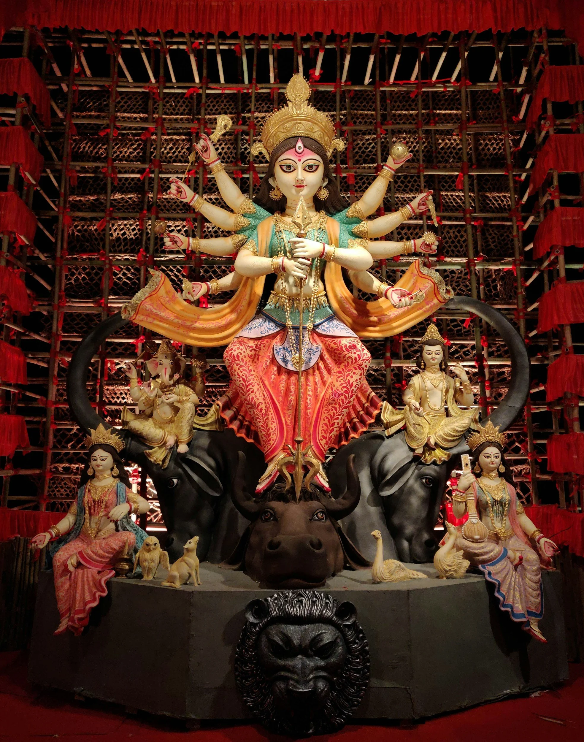 A colorful Hindu sculpture featuring Goddess Durga with multiple arms, riding a lion, accompanied by other deities and animals, set against a red backdrop.