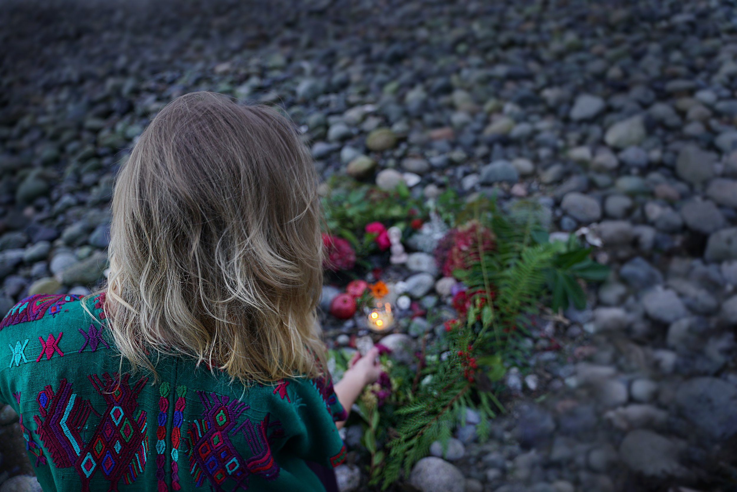 A woman with blonde hair wearing a colorful embroidered shawl, sitting on rocky ground in front of a memorial with flowers and lit candles.