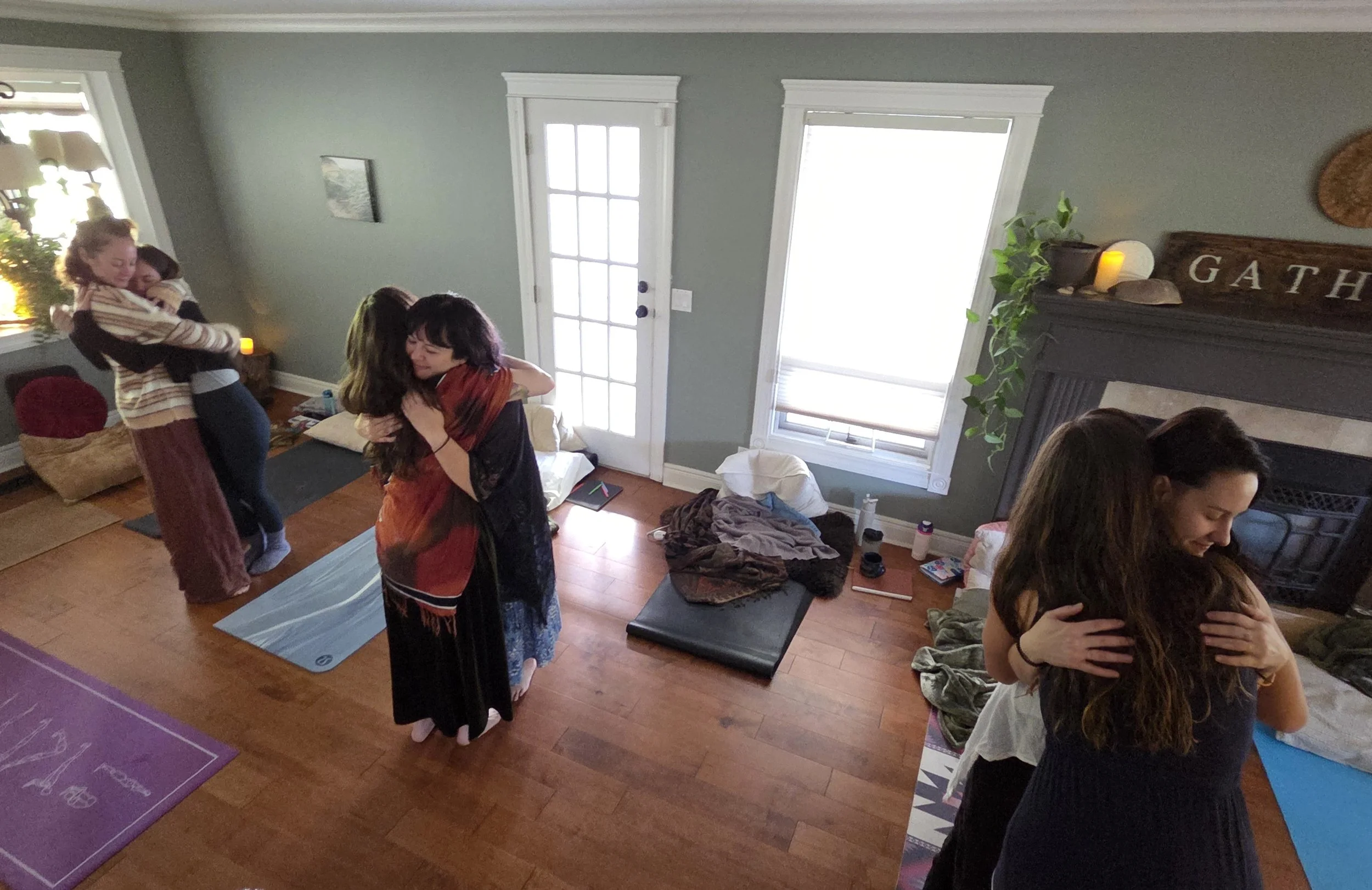Group of women hugging and smiling in a cozy living room, some on yoga mats, with a fireplace, plants, and large windows.