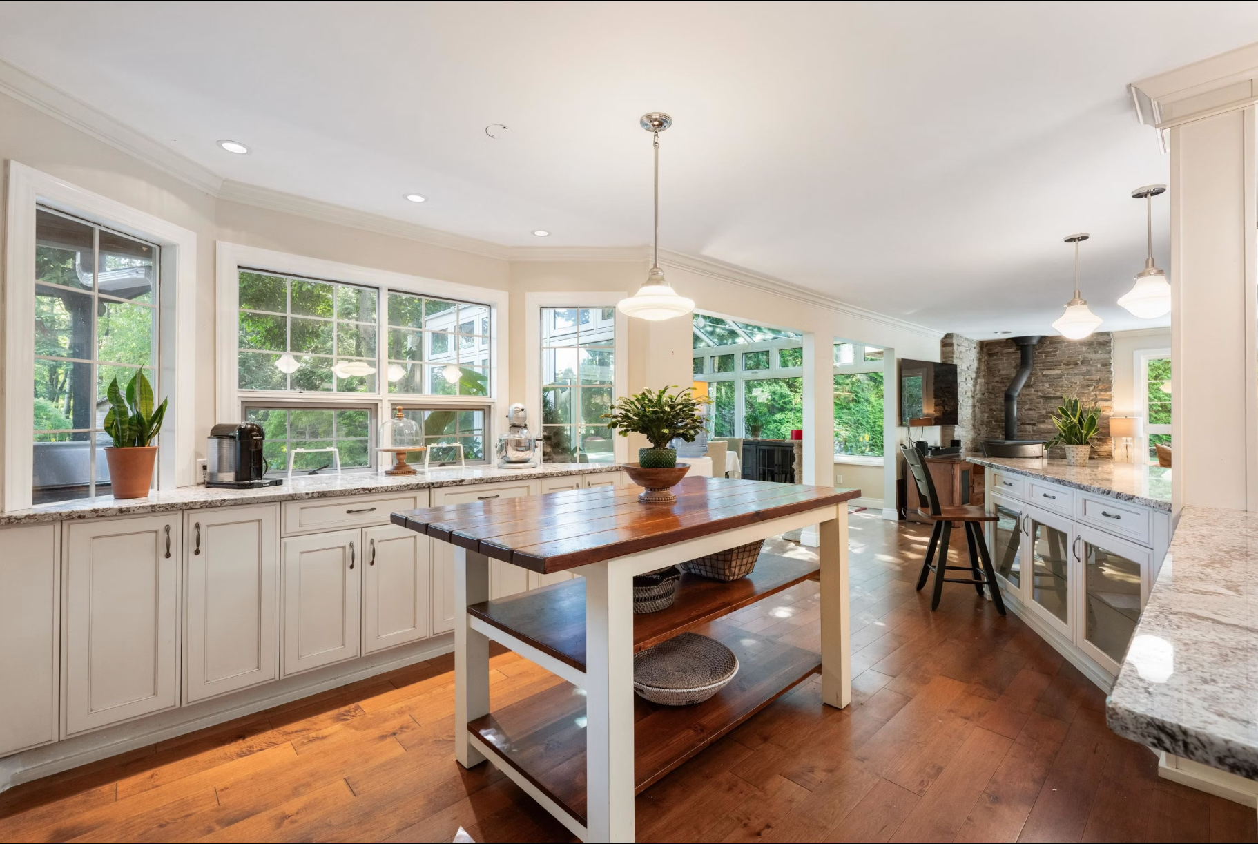Bright, spacious kitchen with white cabinets, granite countertops, large windows overlooking greenery, a wooden kitchen island with a potted plant, and a stone fireplace in the background.