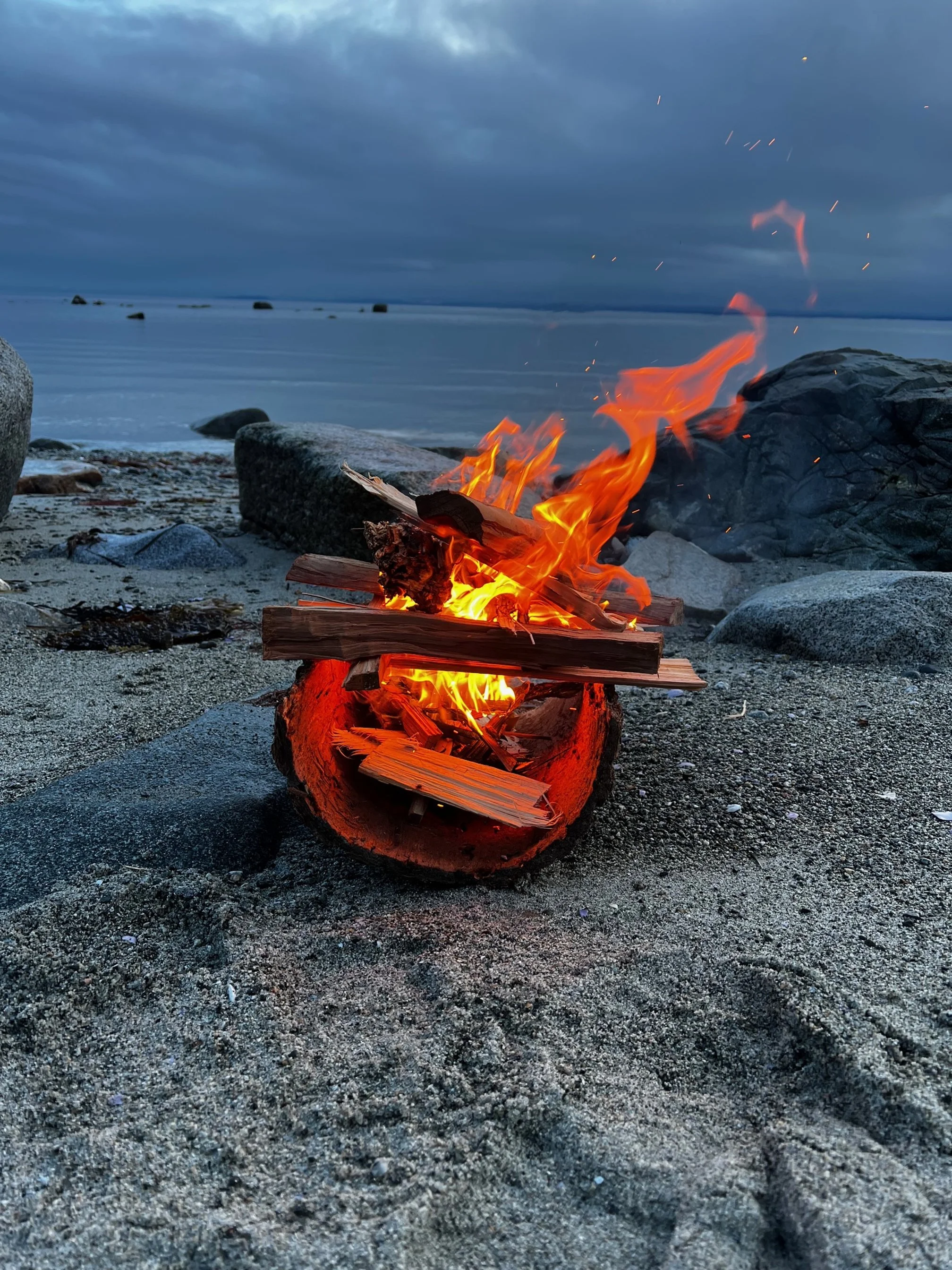 A small campfire burning on a sandy beach near rocks, with a body of water and a cloudy sky in the background.