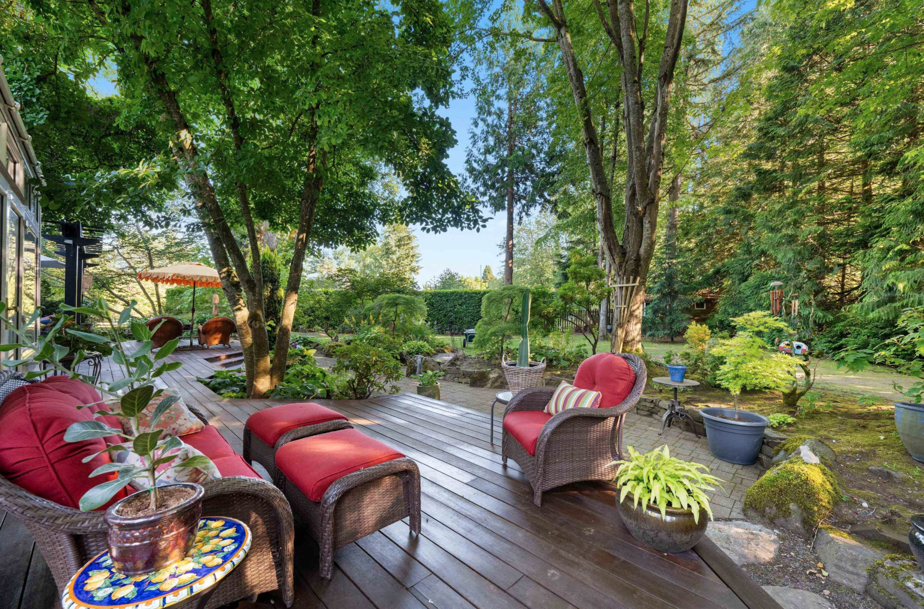 Outdoor patio with wicker furniture, including red cushioned chairs and an ottoman, surrounded by lush green trees and potted plants, under a bright blue sky.