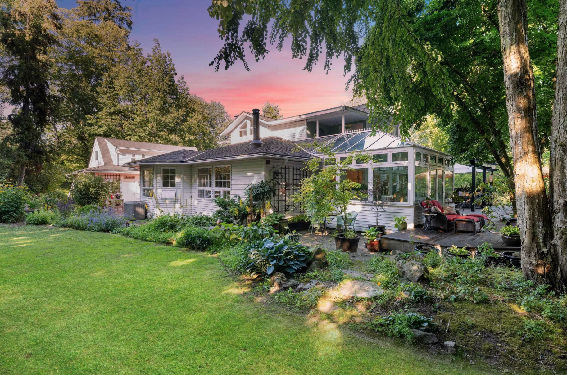 A backyard with a grassy lawn, trees, and a white house with a glass sunroom and outdoor seating area, during sunset.