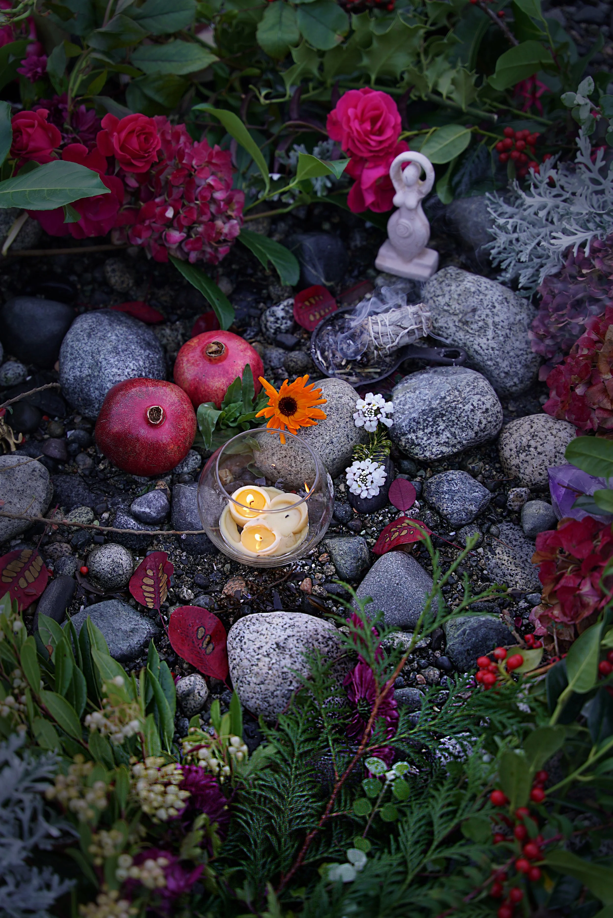 A memorial setup with candles, flowers, apples, rocks, and small statues arranged on the ground among green plants and colorful flowers.