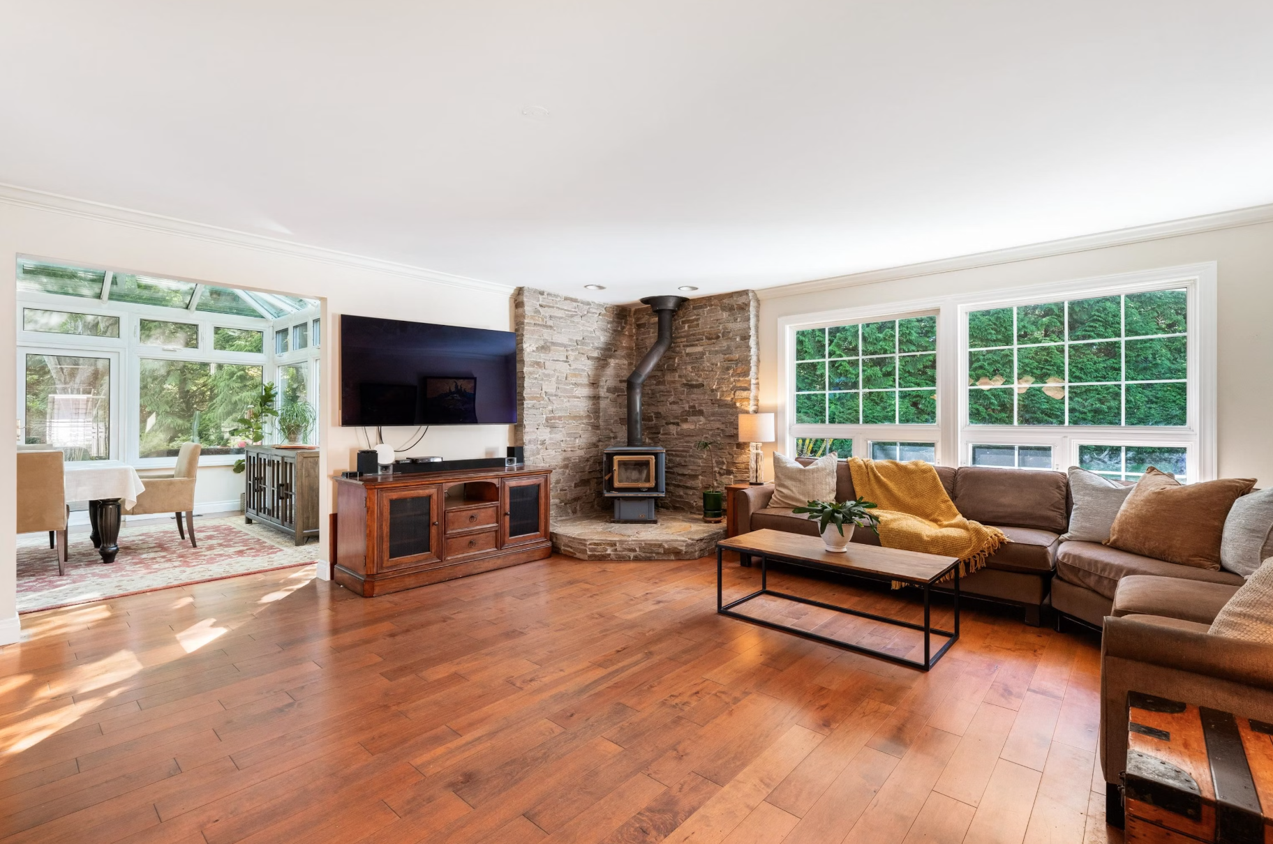 Living room with brown sectional sofa, large windows with green trees outside, a TV on a wooden stand, a fireplace with stone surround and black pipe, a coffee table with a plant, and a lamp beside the sofa.