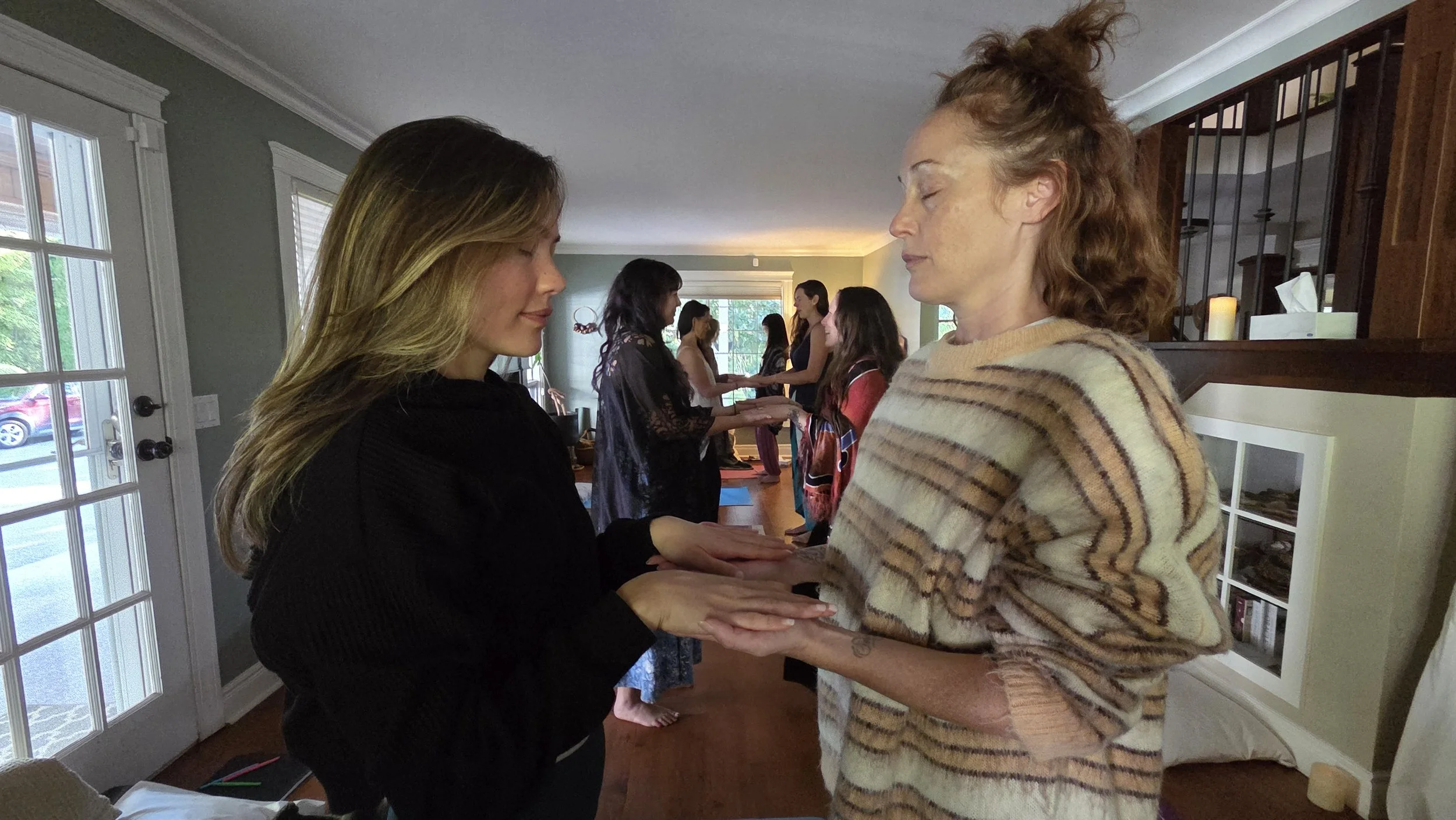 Two women are holding hands face to face, eyes closed, participating in a ritual or ceremony, with more women in the background also involved in similar interactions inside a house.