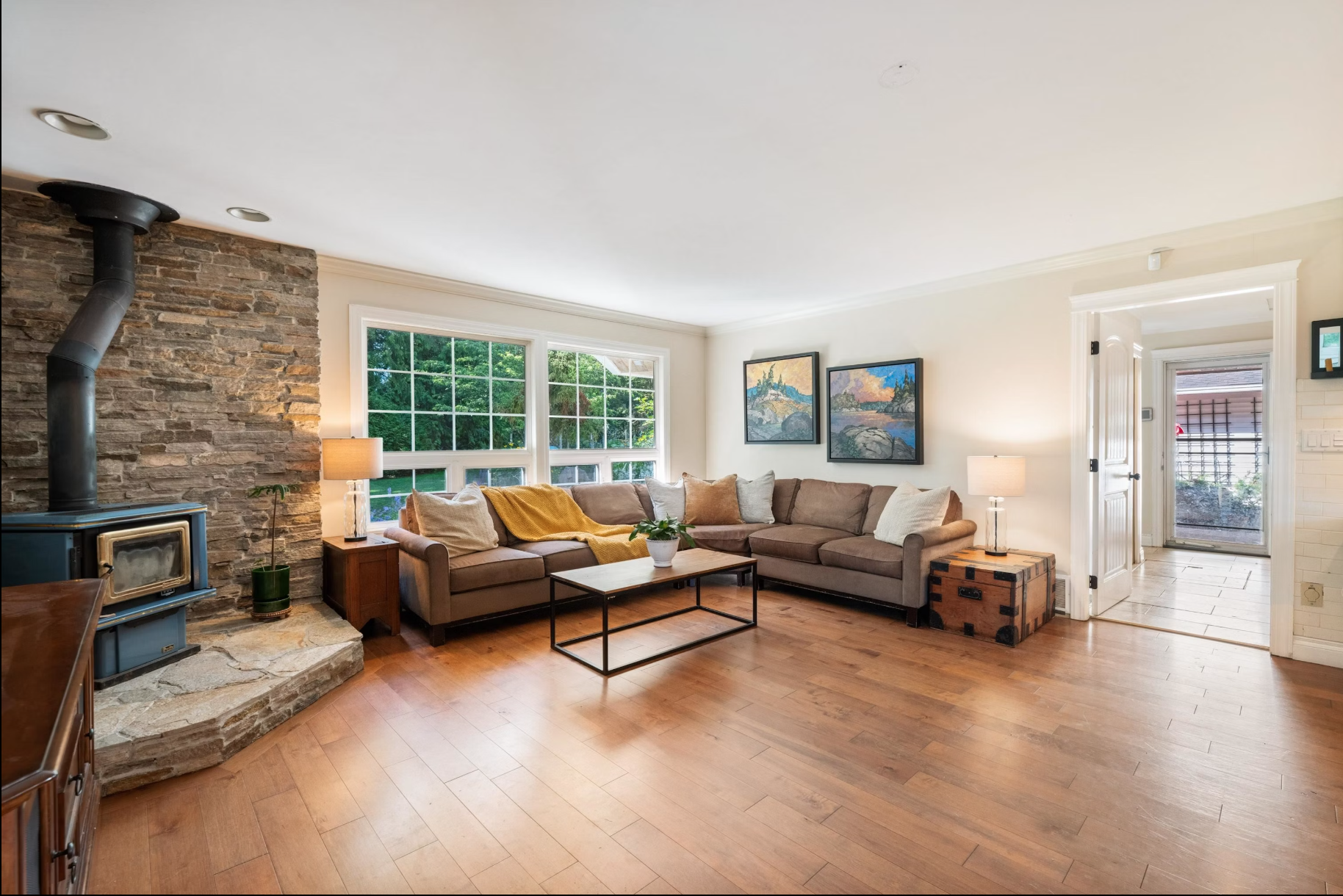 Living room with stone fireplace, beige sectional sofa with pillows and yellow blanket, wooden floor, large window, wall art, side tables with lamps, and an entryway with tiled floor leading outside.