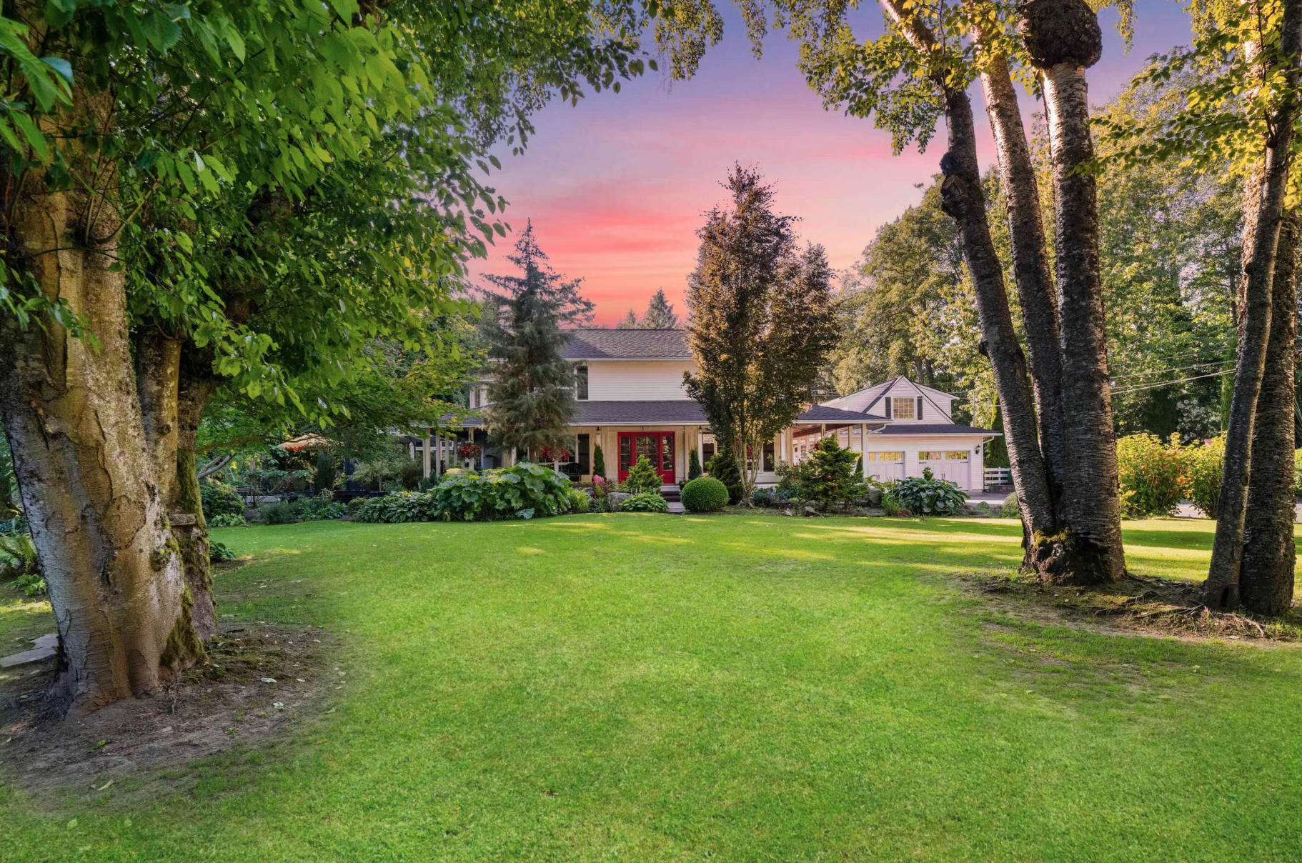 A spacious backyard with lush green grass, large mature trees, and a house with a porch and a garage in the background at sunset.