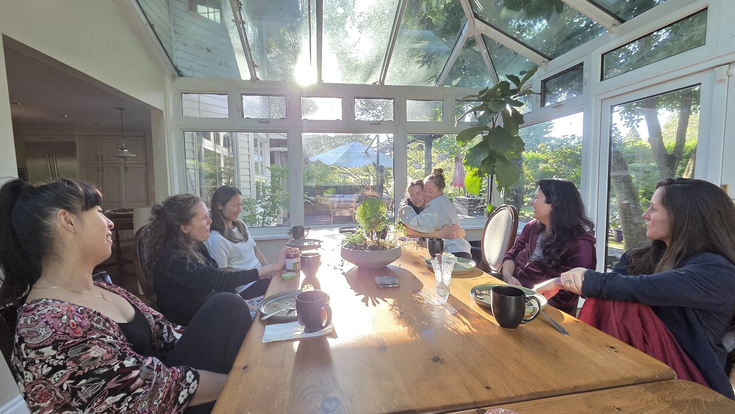 Several women sitting around a wooden dining table in a sunlit sunroom, engaged in conversation and hugging, with greenery visible outside through large windows.