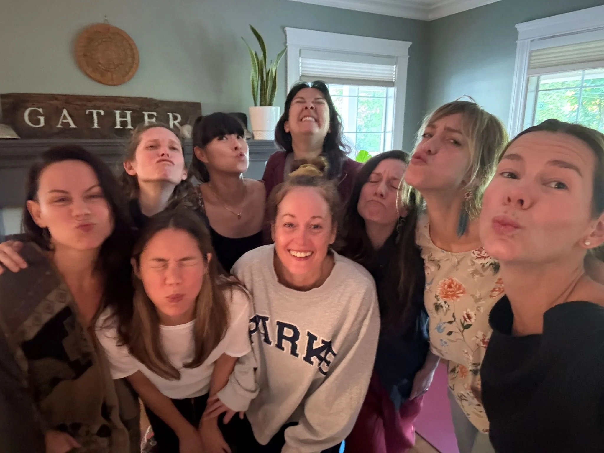 Group of nine women making silly faces in a cozy living room with a window, couch, and decorative sign that says 'Gather' in the background.