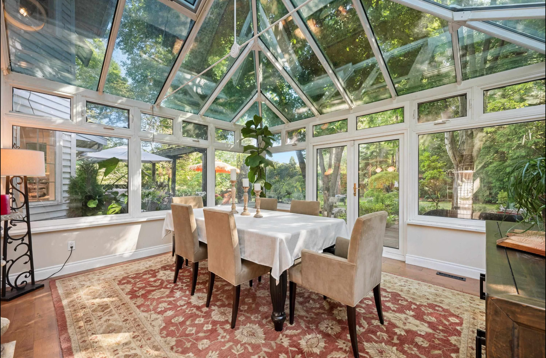Sunroom with glass ceiling and windows, beige dining chairs, white tablecloth, candelabras, and view of green garden outside.