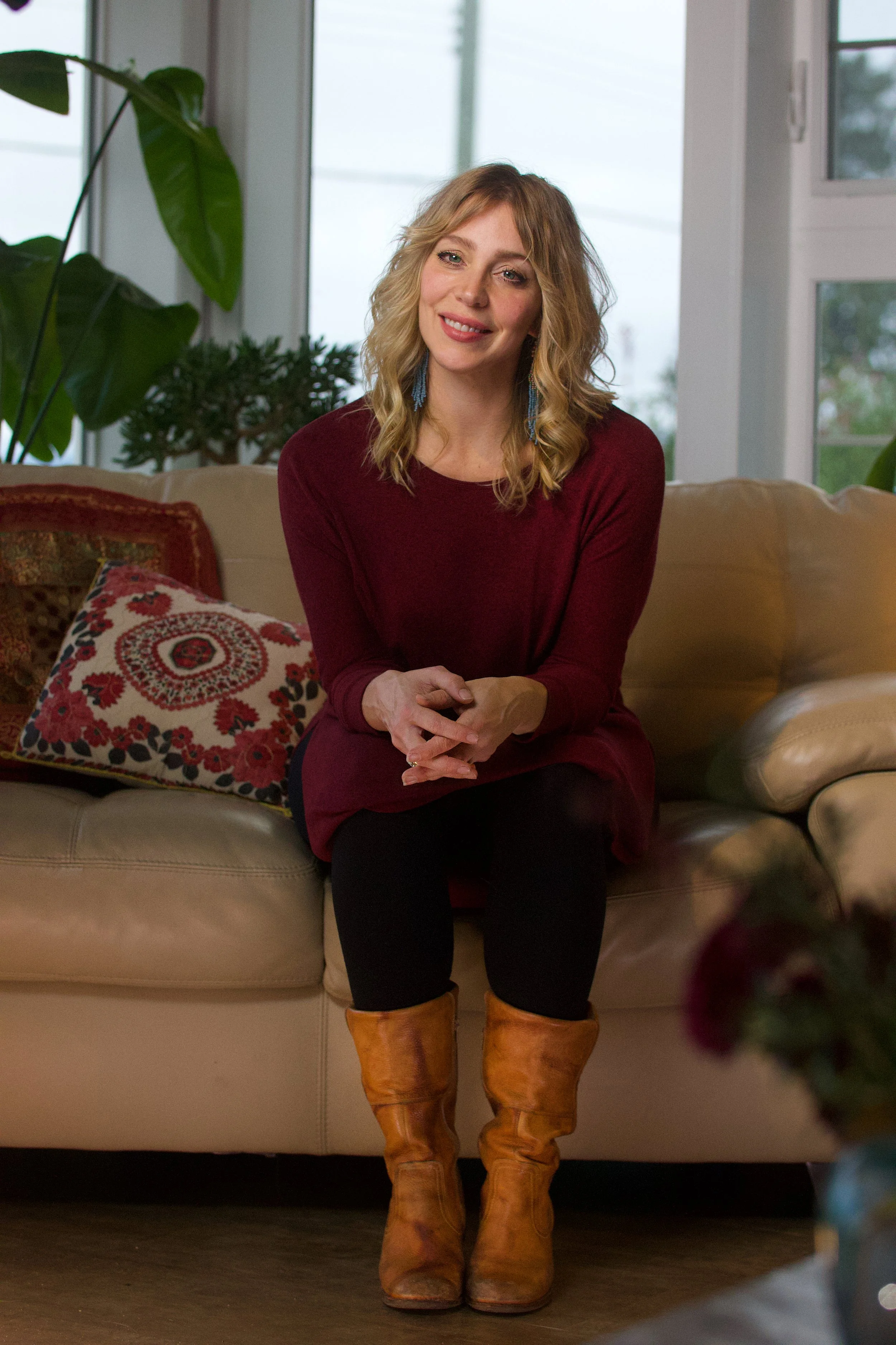 A woman with wavy blonde hair wearing a maroon top and brown boots sitting on a beige couch in a room with large windows and green plants.
