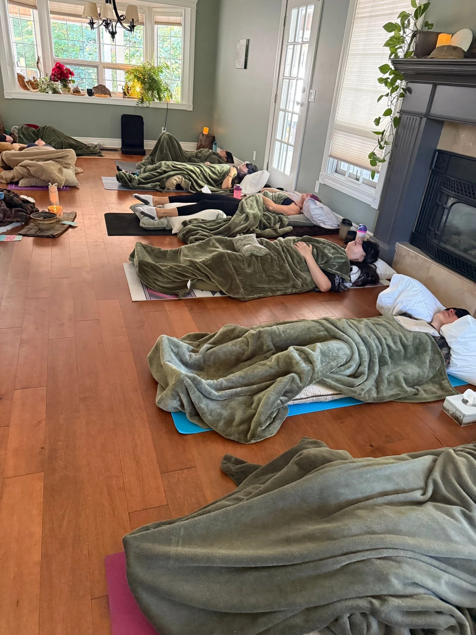 People lying on yoga mats covered with green blankets in a bright living room with large windows, a fireplace, and wooden floors, participating in a group relaxation or meditation session.