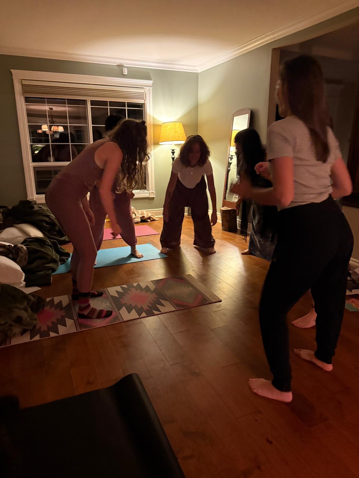 Group of women doing yoga or stretching exercises together in a living room at night with warm lighting, a mirror, and large window.
