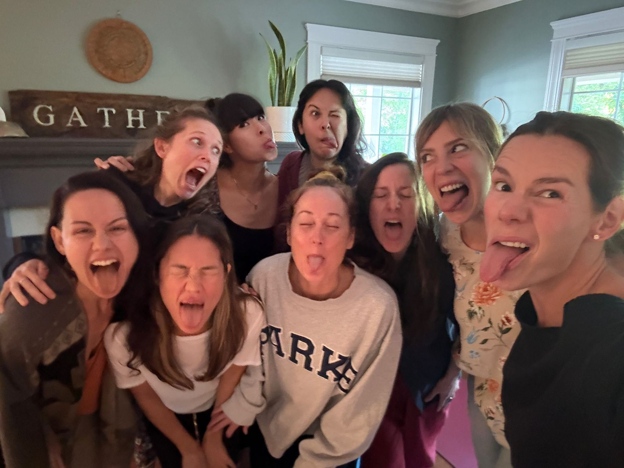 Ten women posing together indoors, making silly faces with tongues out and eyes squinted, in a casual gathering or house setting.