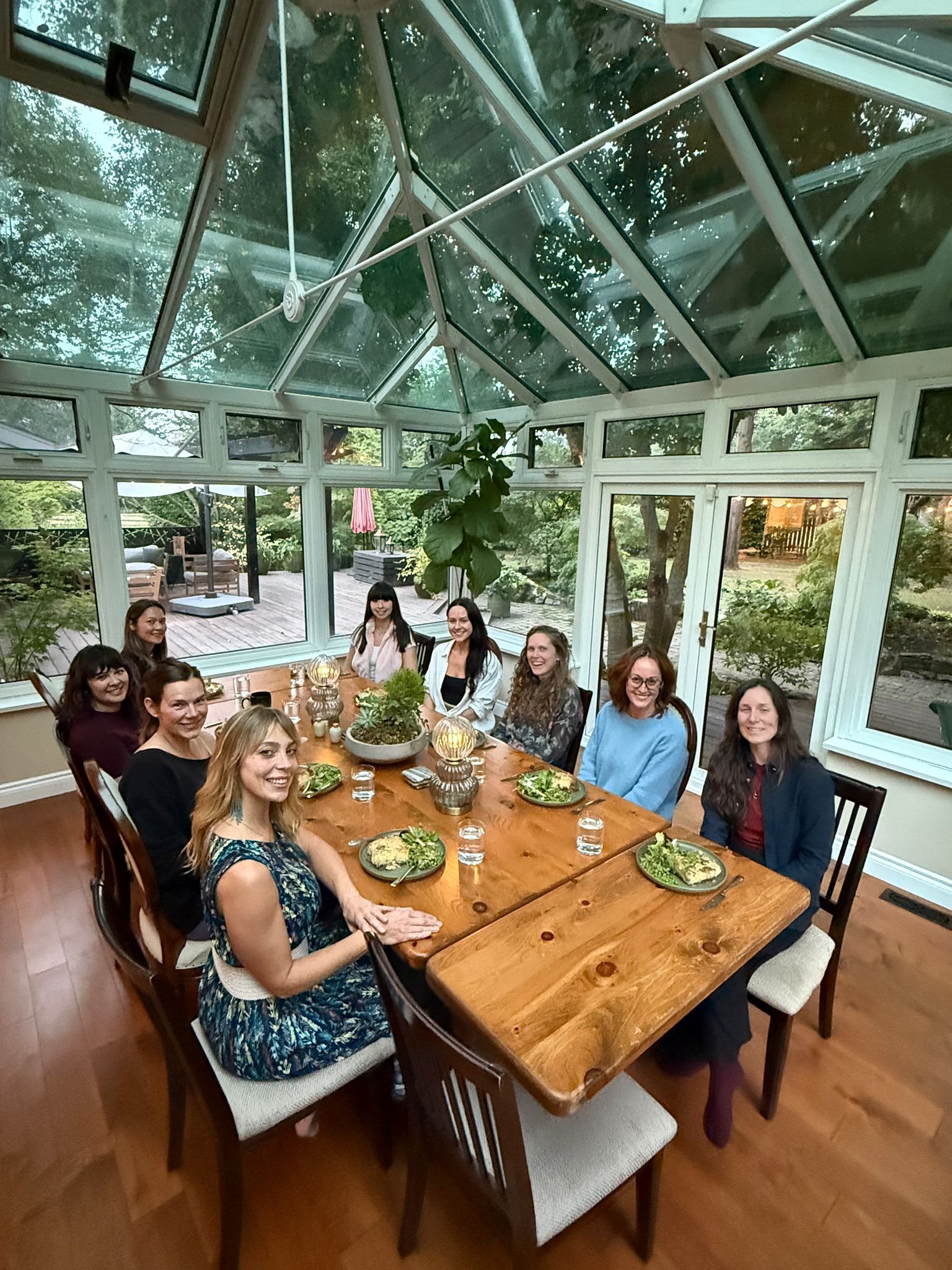 A group of nine women sitting at a wooden dining table inside a glass conservatory, smiling at the camera.