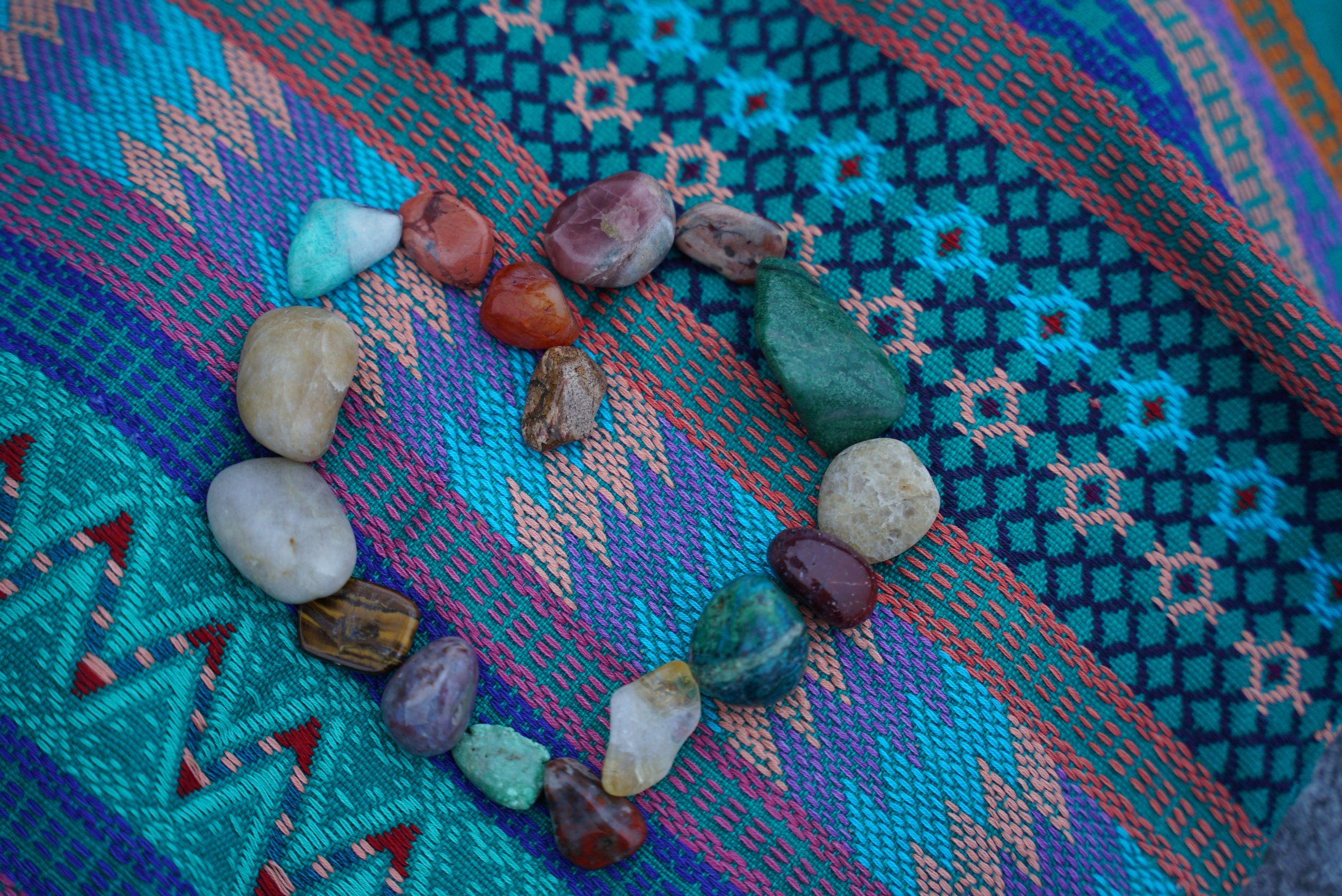 Colorful woven fabric with a semi-circle arrangement of various small, polished stones, including green, brown, red, purple, and white stones.