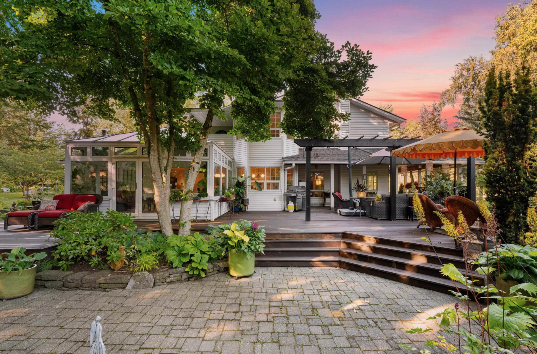 A spacious backyard patio at sunset featuring a large wooden deck with outdoor furniture, a gazebo with an umbrella, potted plants, a tree, and a white house in the background.