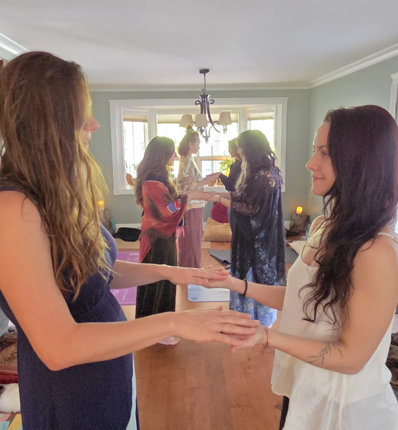 Group of women participating in a yoga class or meditation session in a living room, facing each other with hands extended, in a circle formation.