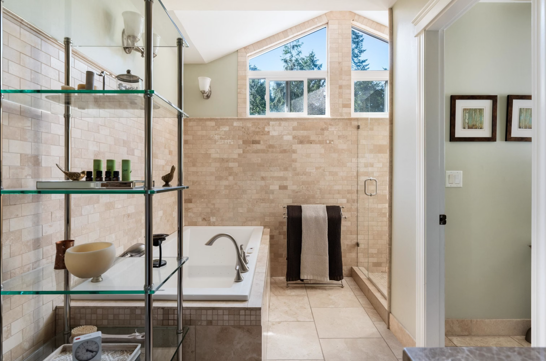 Bathroom with a bathtub, glass shelves with decorative items, tiled walls, a window with trees outside, and a towel rack with two towels.