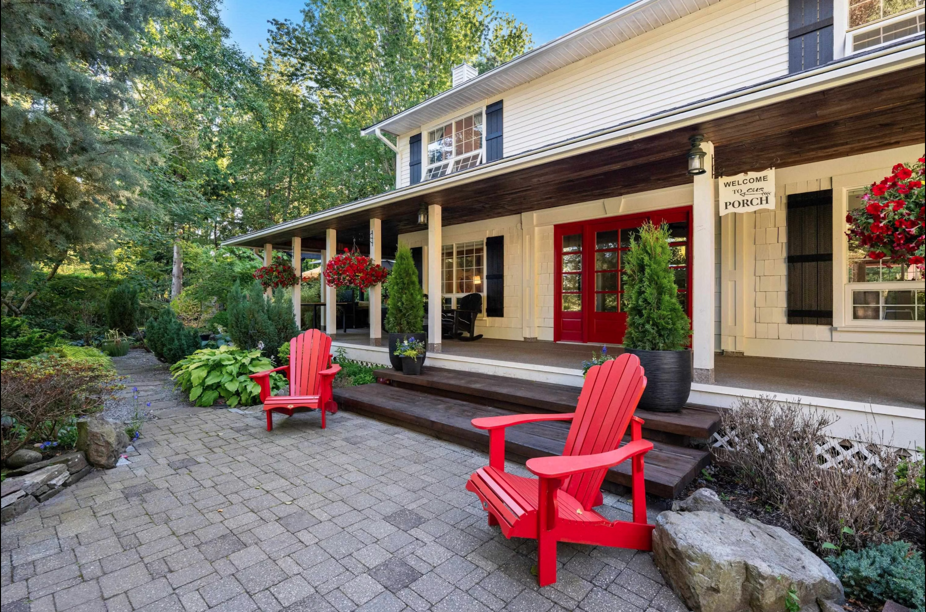 Front porch of a house with red double doors, two red Adirondack chairs on a stone patio, potted plants, hanging flower baskets, and a garden with shrubs and trees.