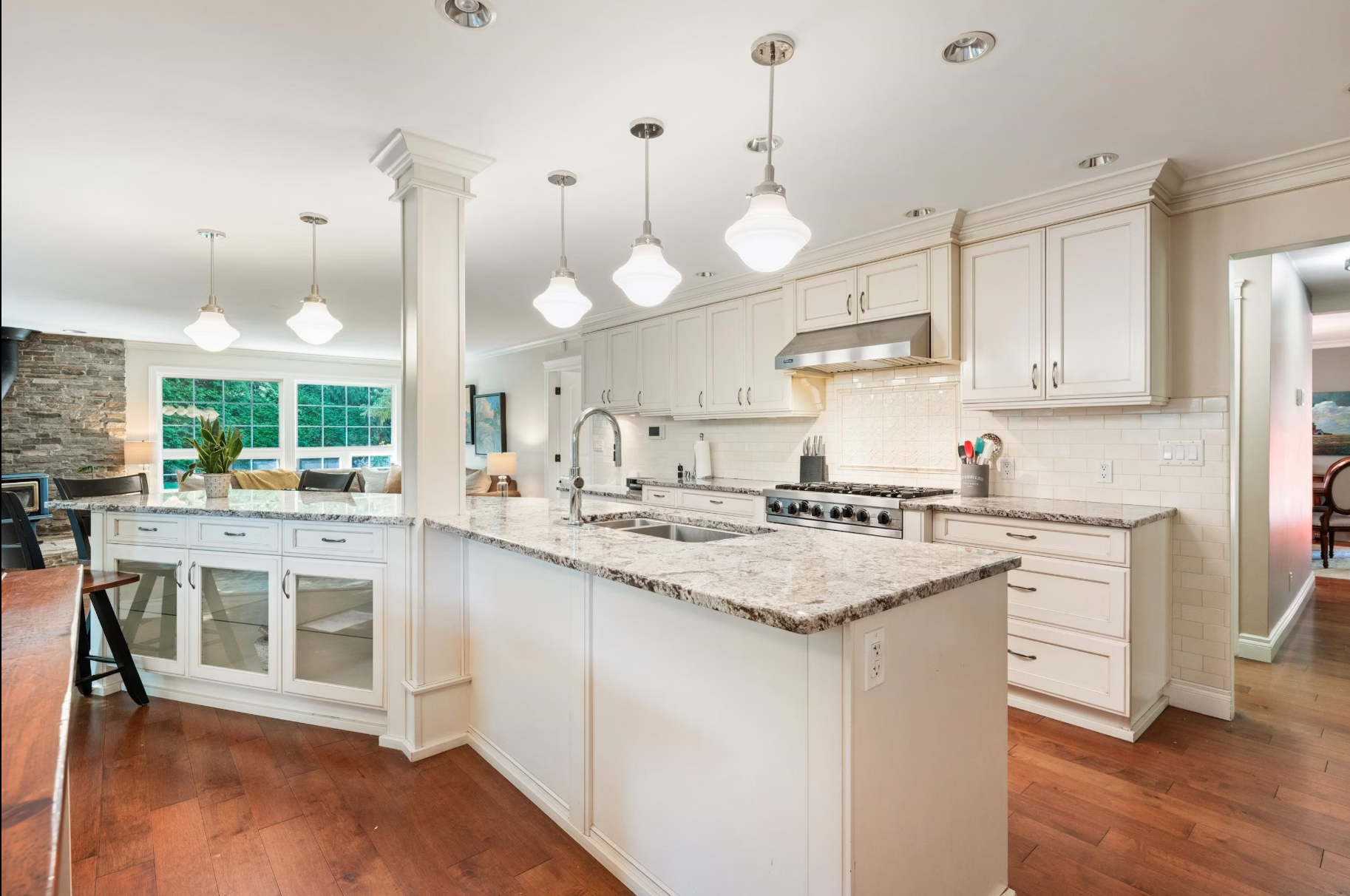 Modern kitchen with white cabinets, granite countertops, stainless steel appliances, pendant lighting, hardwood floors, and a view of the living area with large window and stone wall.