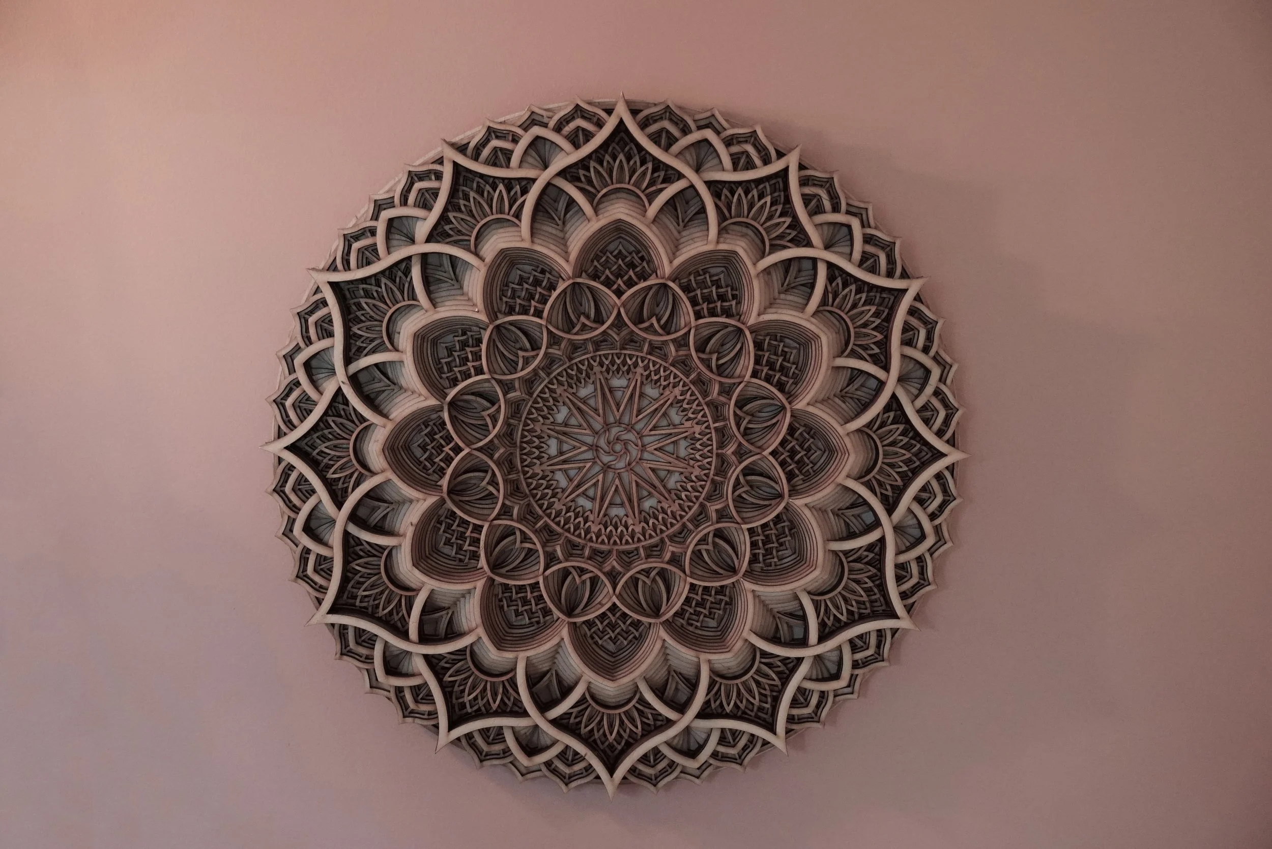 A circular paper or cardboard decoration with intricate geometric patterns and layered designs, mounted on a plain pink wall.