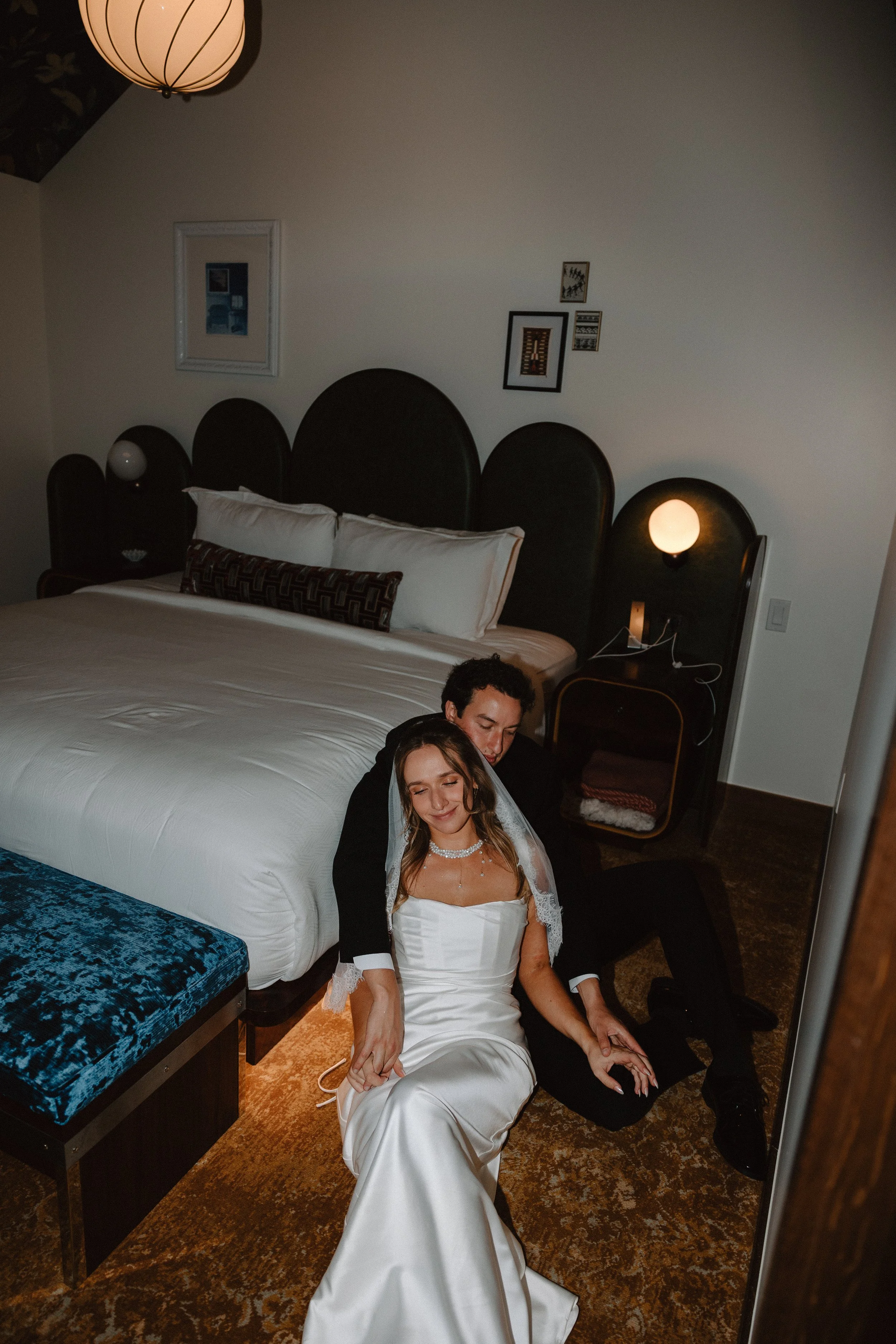 A bride and groom sitting on the floor of a hotel room in front of a large bed, smiling with their eyes closed. The bride is wearing a white satin wedding dress and veil, while the groom is dressed in black.