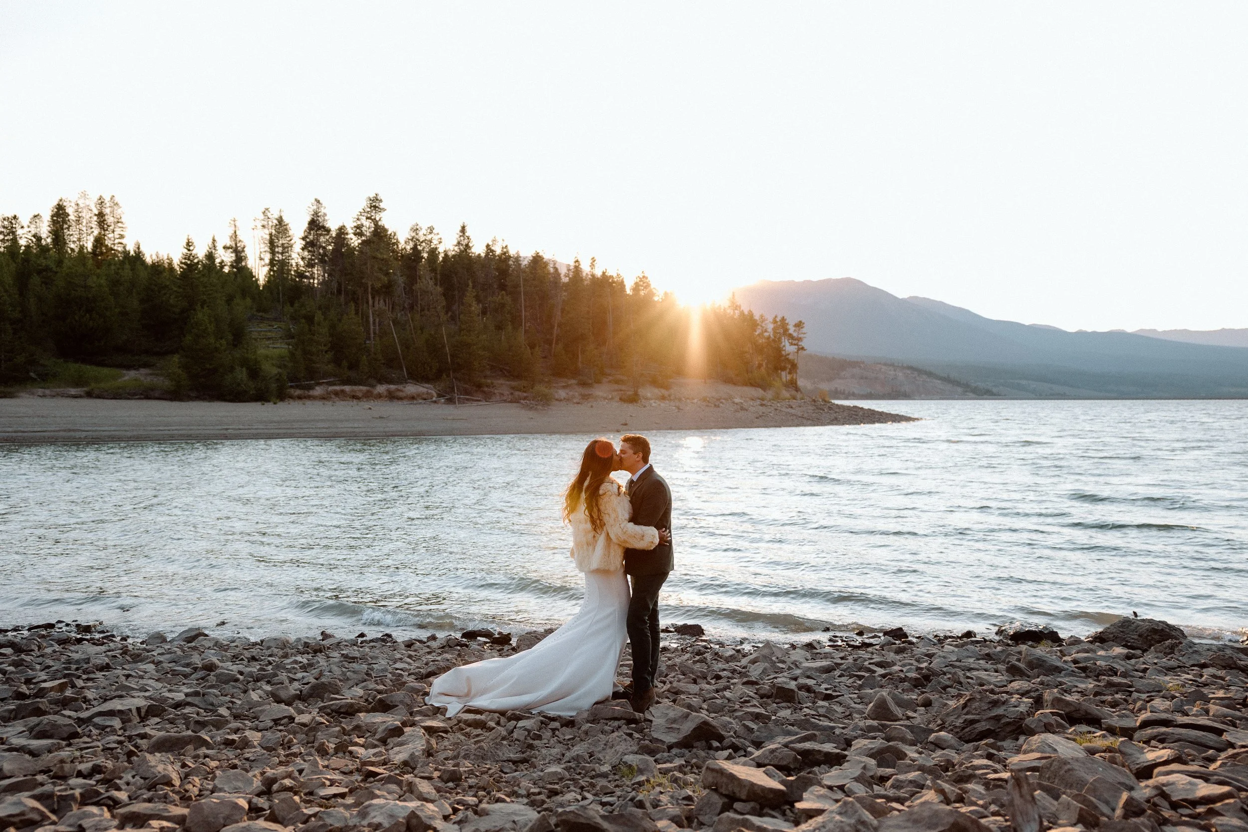 Couple in wedding attire kissing on rocky beach at sunset with lake, trees, and mountains in background.