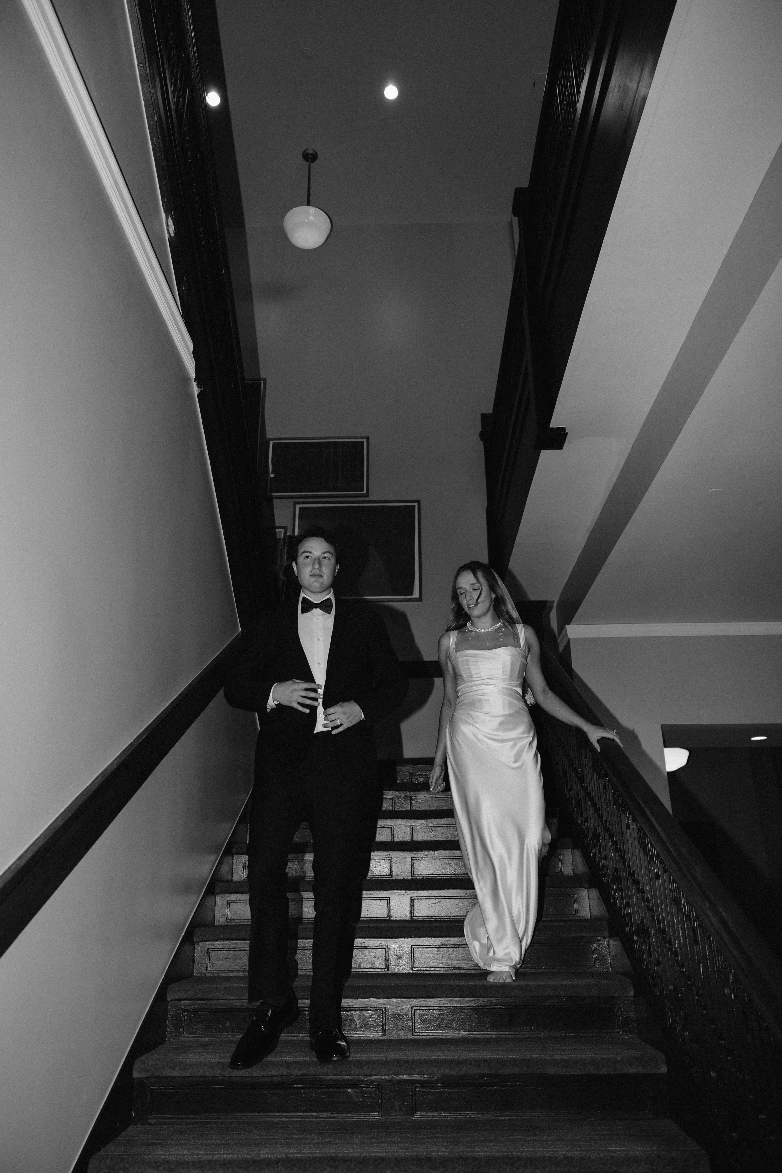 A young man in a tuxedo and a young woman in a satin gown are walking down a staircase in an interior setting.