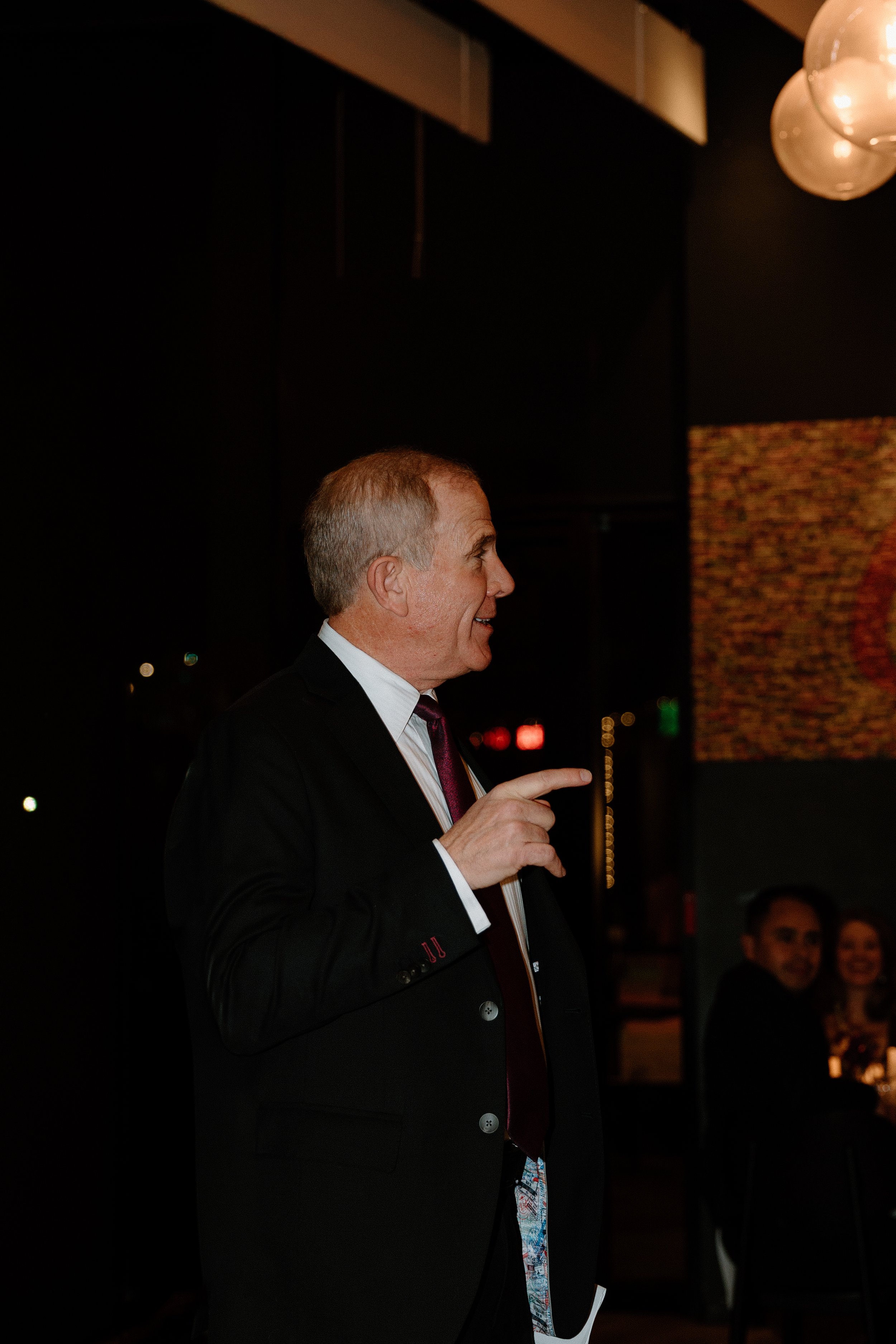 A middle-aged man in a black suit and tie pointing and smiling at an indoor event with other people seated in the background.